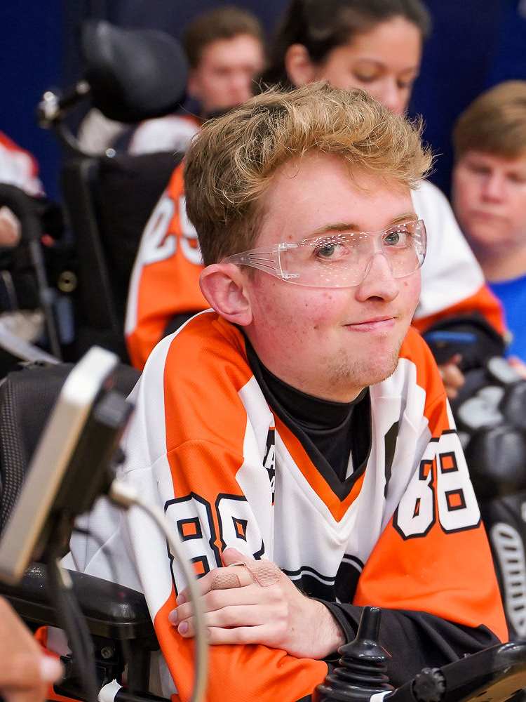 Liam Miller, a young man wearing safety glasses and an orange and black hockey jersey, smiles while seated in wheelchair