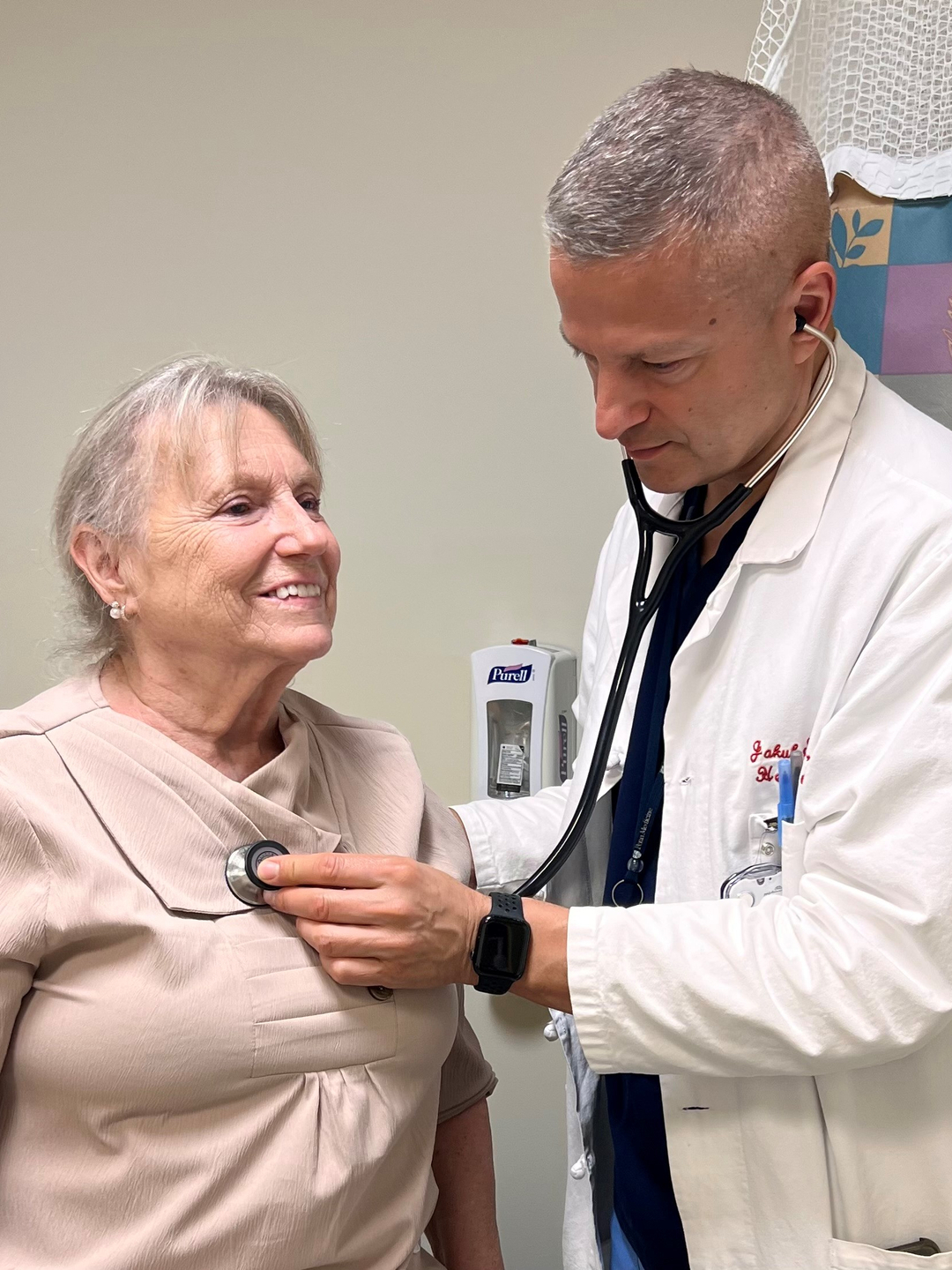 Jakub Svoboda, MD, holds a stethoscope to the heart of Jane Gesemyer, a woman in her 70s, who is smiling