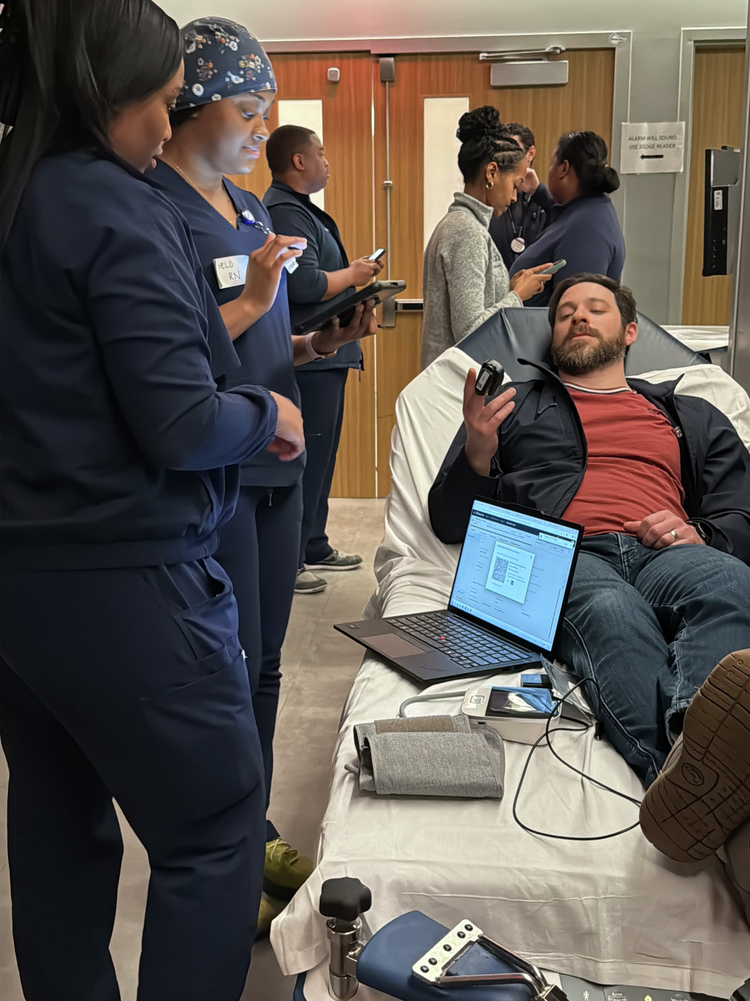 Two nurses talk to a patient who is laying on a gurney in a hallway with a pulse oximeter attached to his index finger. An open laptop and a blood pressure cuff are also on the bed