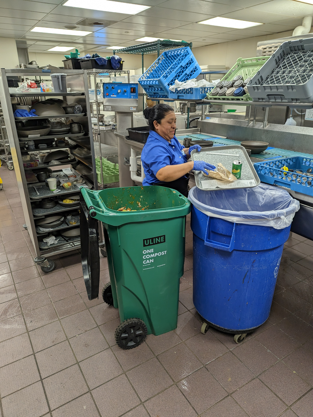 A food services worker in a kitchen tips a tray toward a waste bin