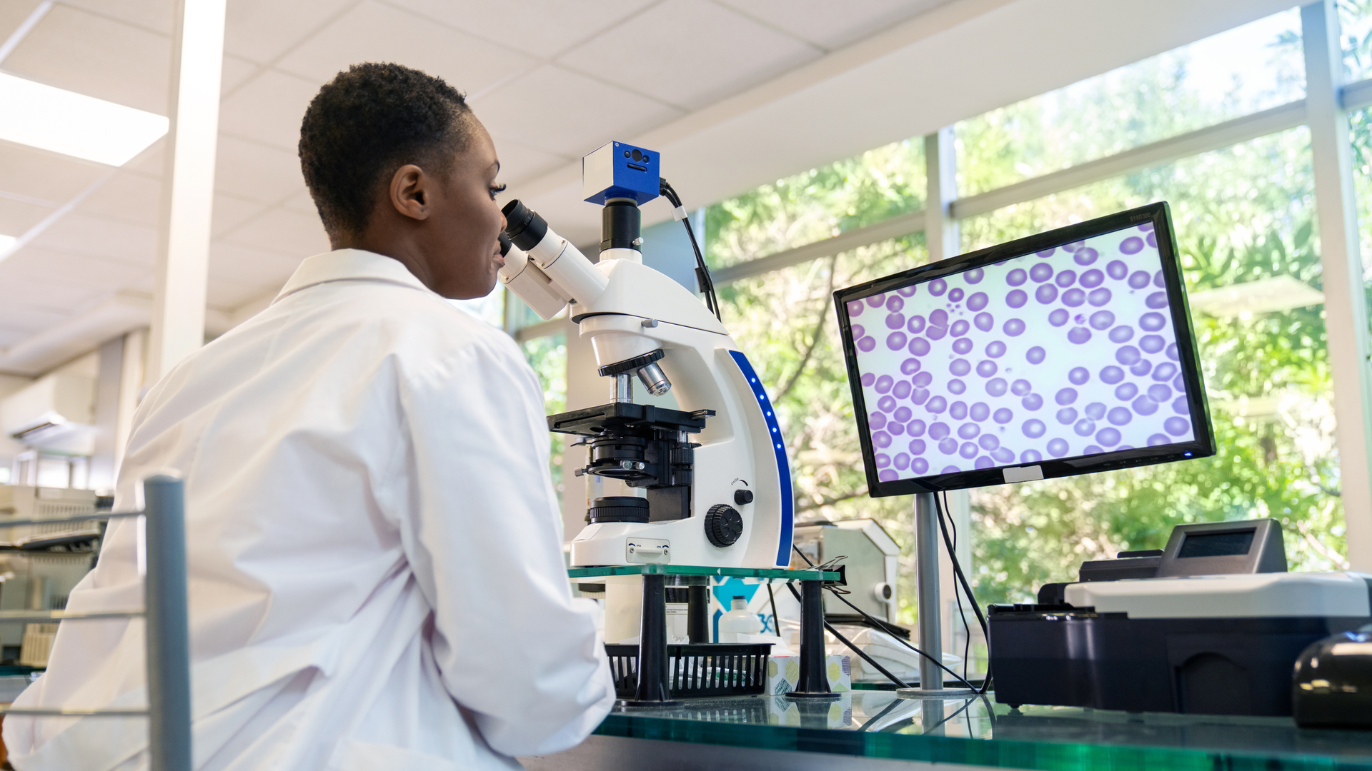 Researcher in lab coat looking at cells through microscope while cells are displayed on monitor