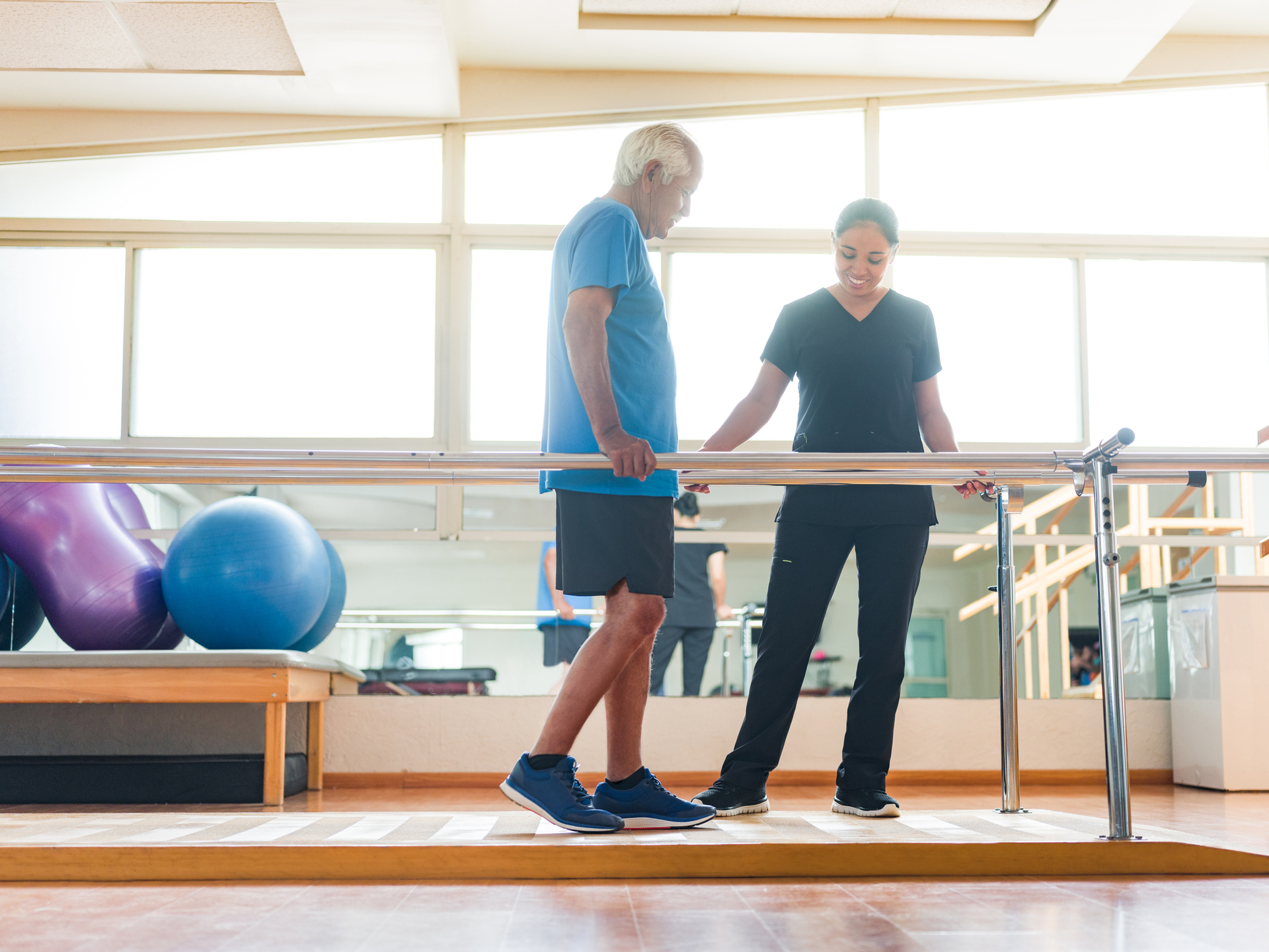 A senior man giving small steps in the parallel bars and physical therapist standing next to him.