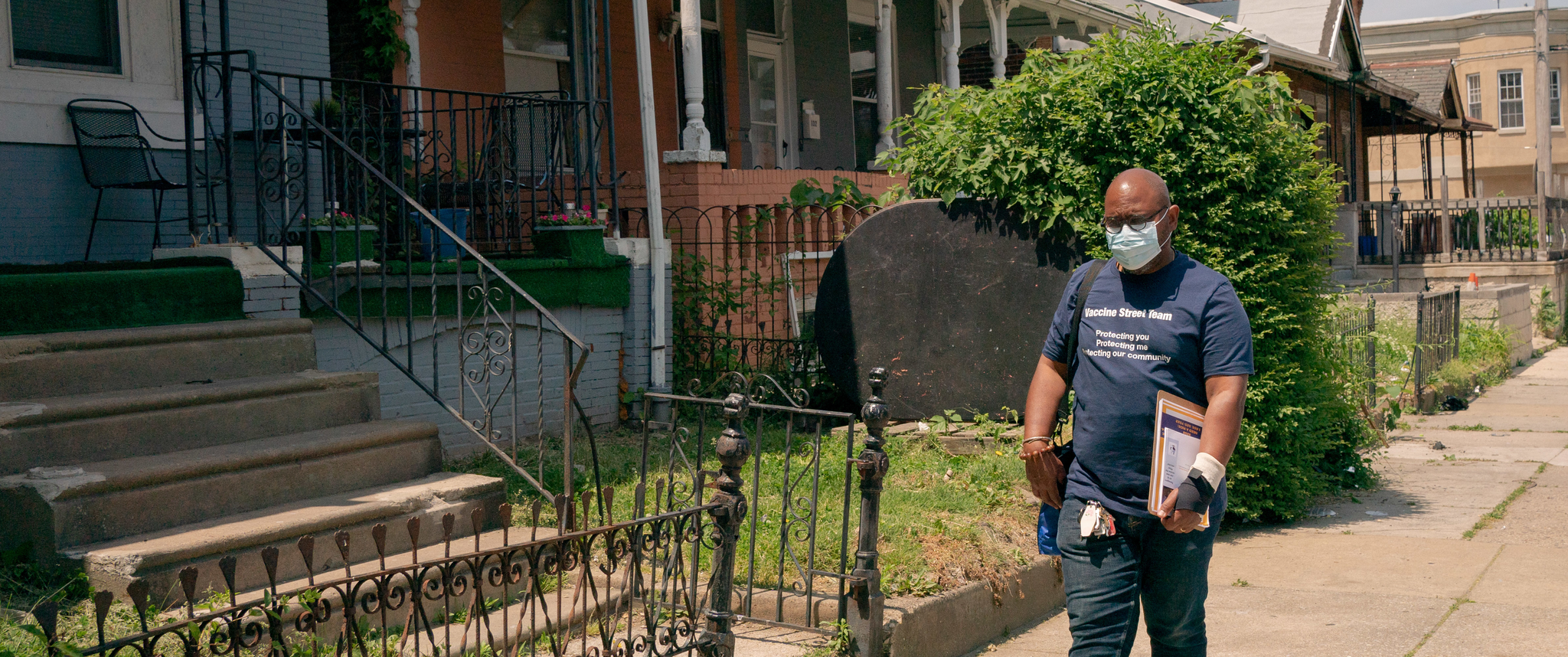 Yuhnis Syndor, wearing a mask and carrying a clipboard, walks on a West Philadelphia residential sidewalk