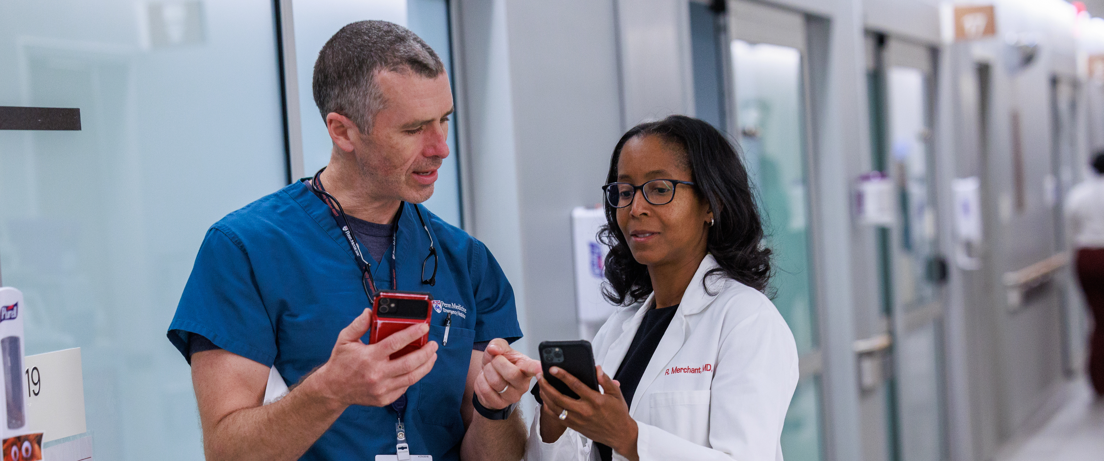  Bryan Walker Lee, MD and Raina Merchant, MD, stand in the emergency department wearing scrubs and a white coat, looking at cell phones