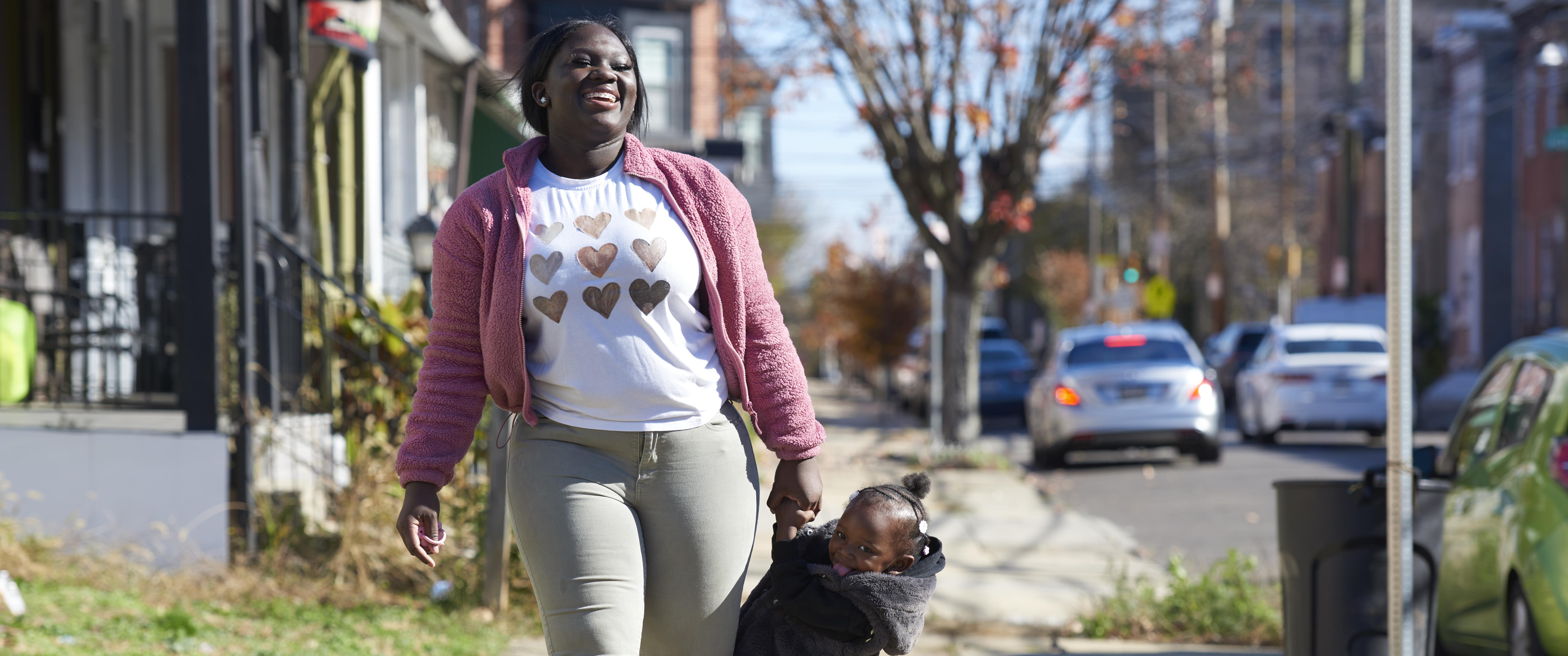 Talicia Williams walks down a city sidewalk, smiling, as her 2-year-old daughter hangs playfully from her arm