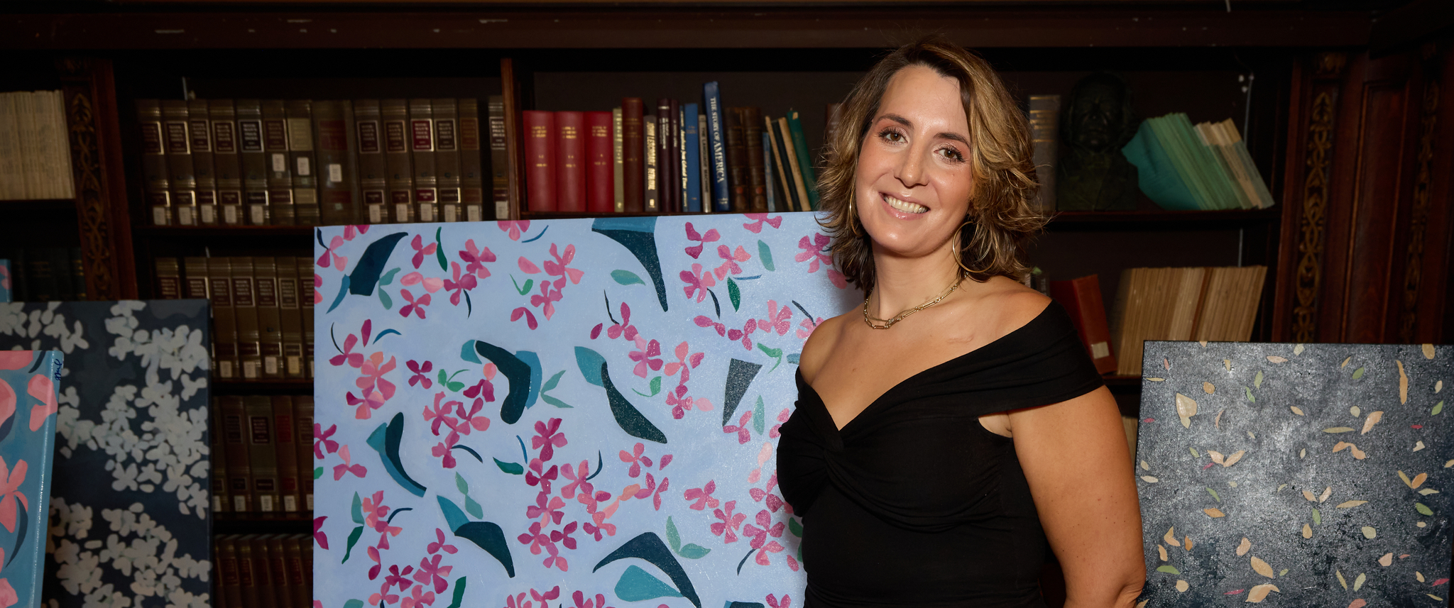 Ginger Mimmo Rohlfing standing in front of her painting, one with pink flowers and blue leaves on a pale blue background, smiling in a black dress