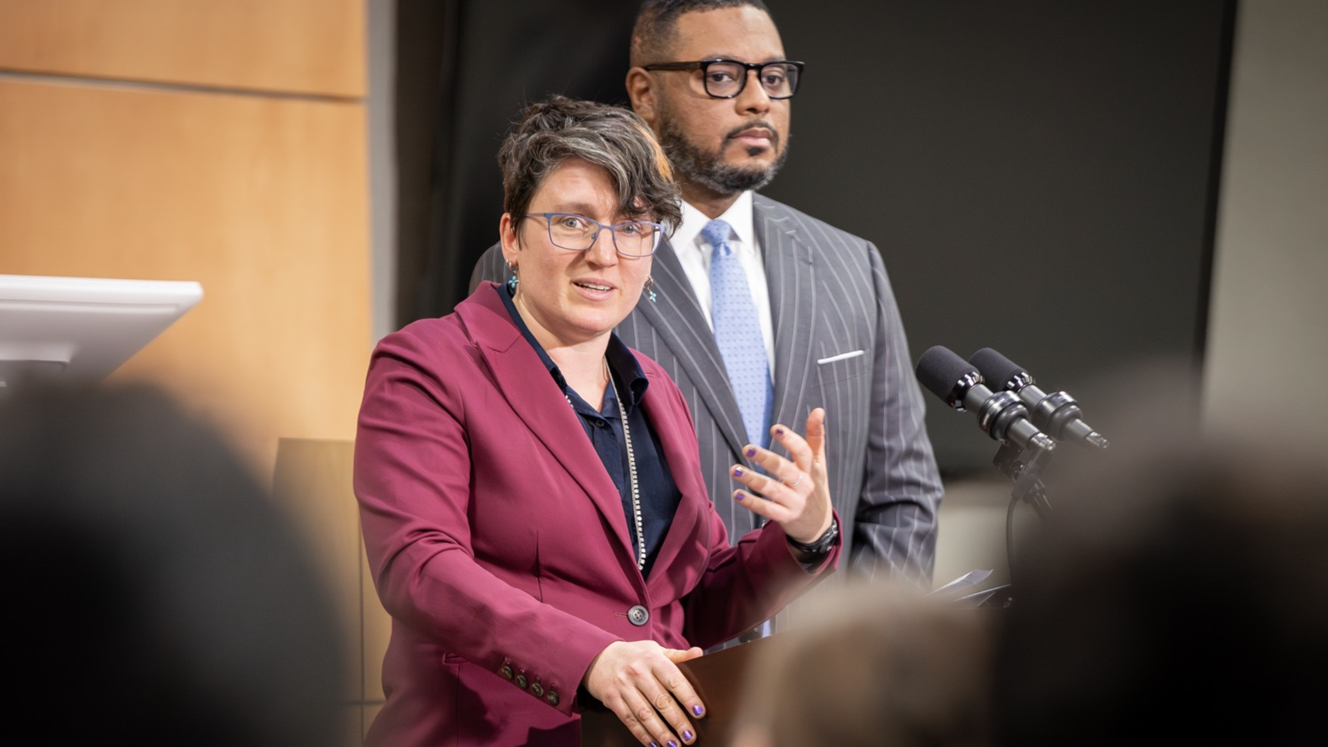 Woman in maroon blazer stands at a podium next to a man in a gray pinstripe suit. 