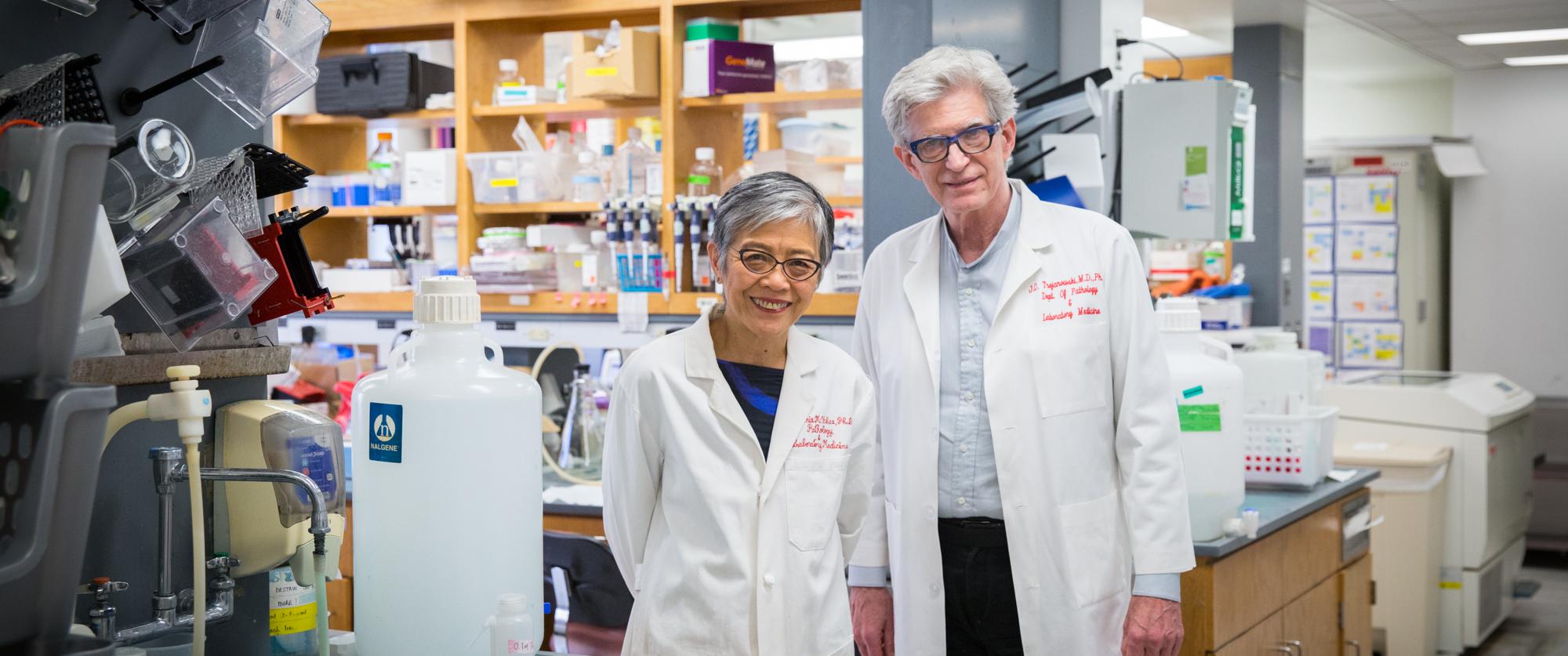 Portrait of Virginia Lee and John Trojanowski standing in a lab wearing white coats