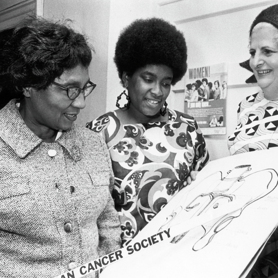 Dickens stands with three other women dressed in 1970s styles, holding an oversized pencil that points to a drawing of fallopian tubes and ovaries inside a pelvic bone.