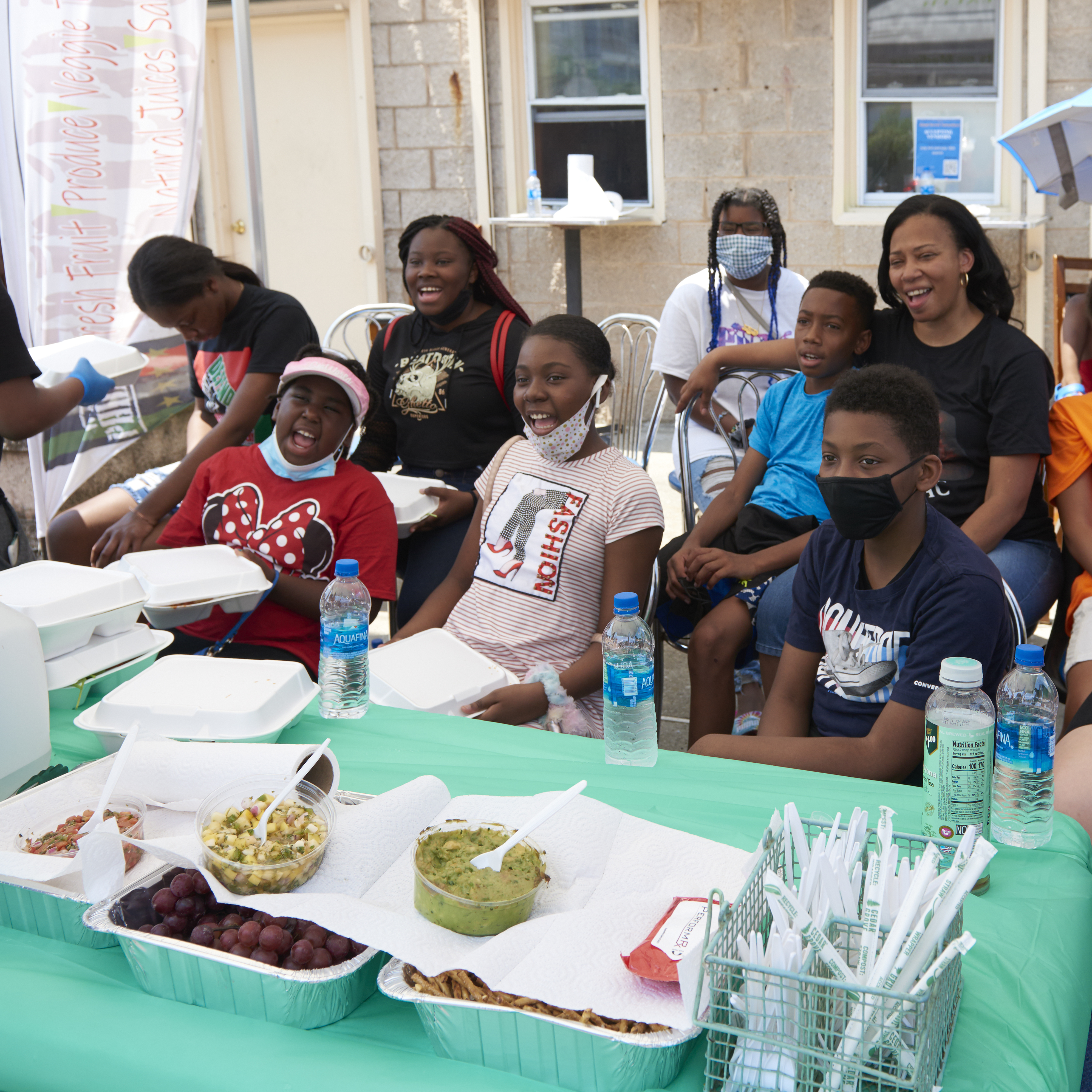 A group of children, teens, and adults seated in folding chairs outdoors smile as a boy hands out food