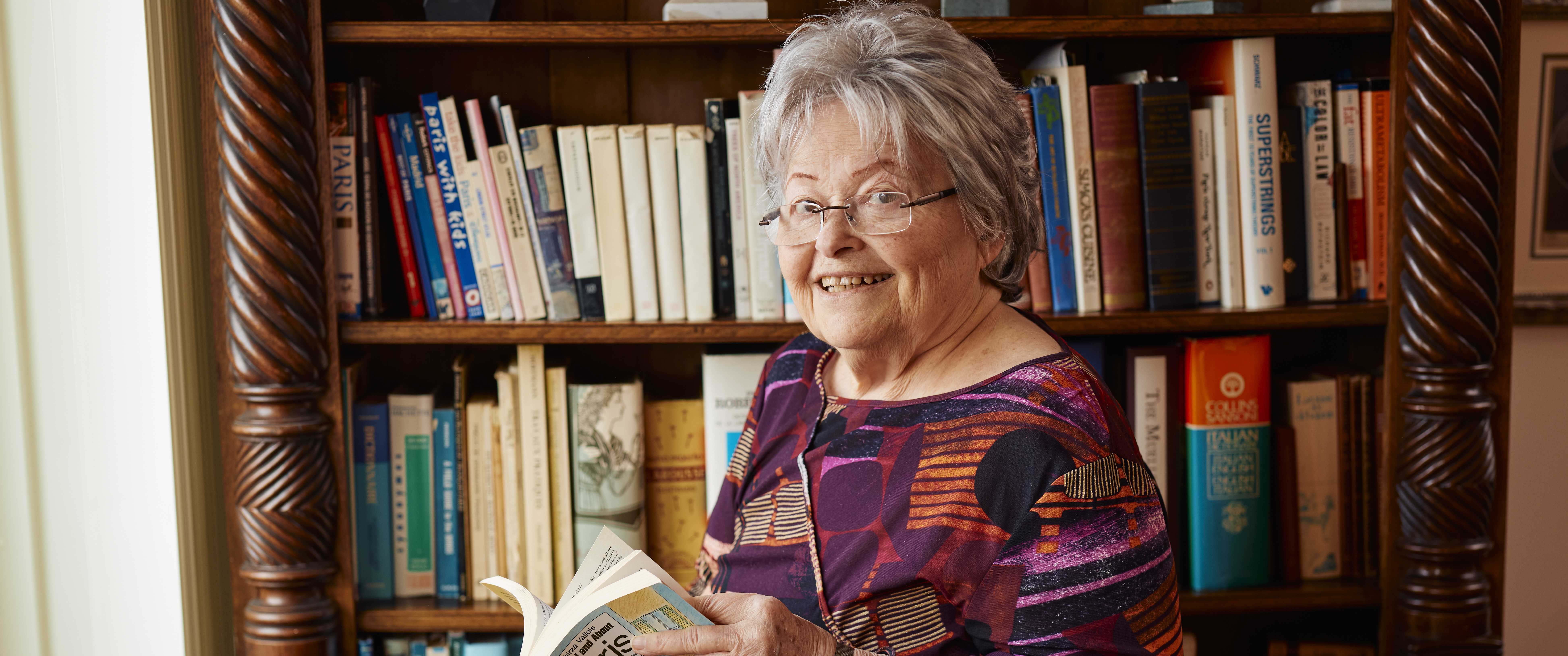 Mimi Reed, smiling, stands in front of a bookcase at home, holding a book about Paris