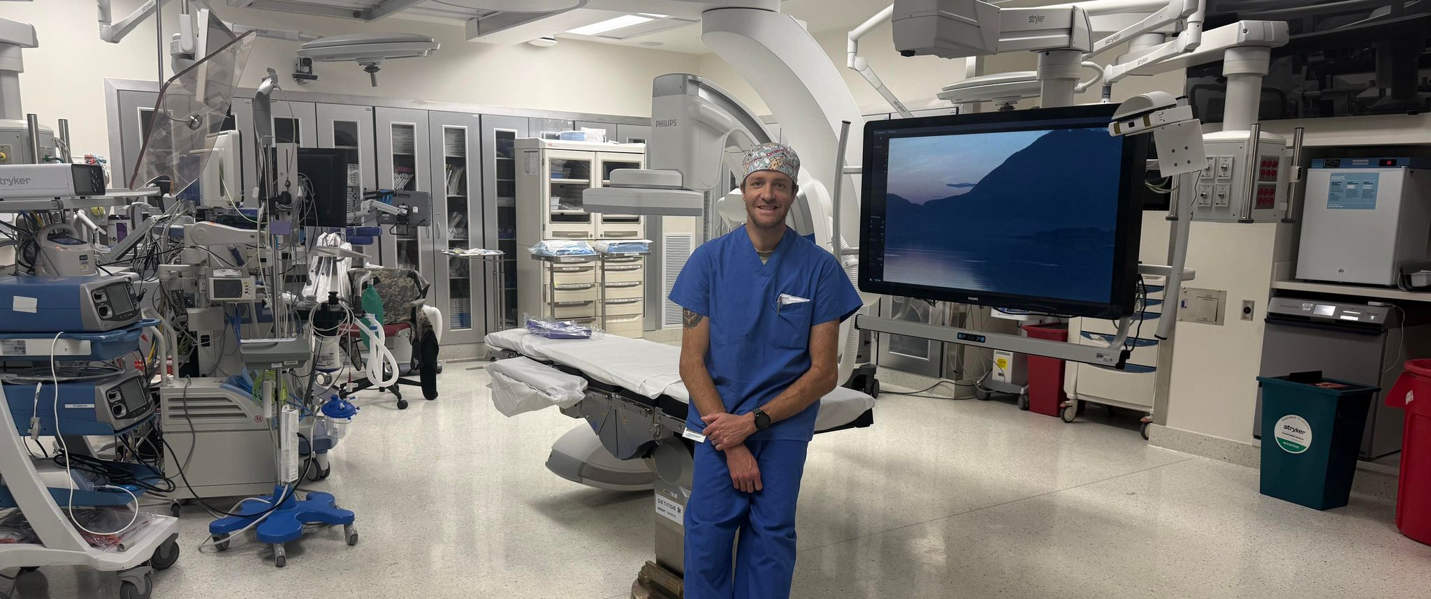 Chris Naimo, wearing scrubs, stands smiling in an empty operating room