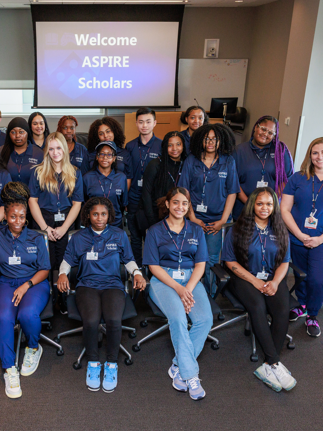 Several dozen young people pose under a screen projecting “Welcome ASPIRE Scholars”