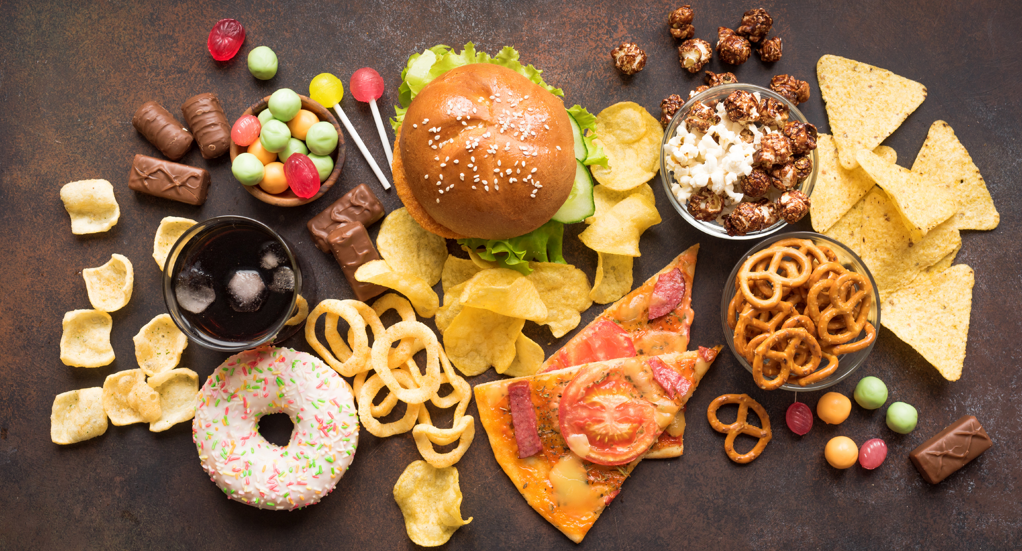 Food on a table, including a burger, pizza slices, chips, and candy