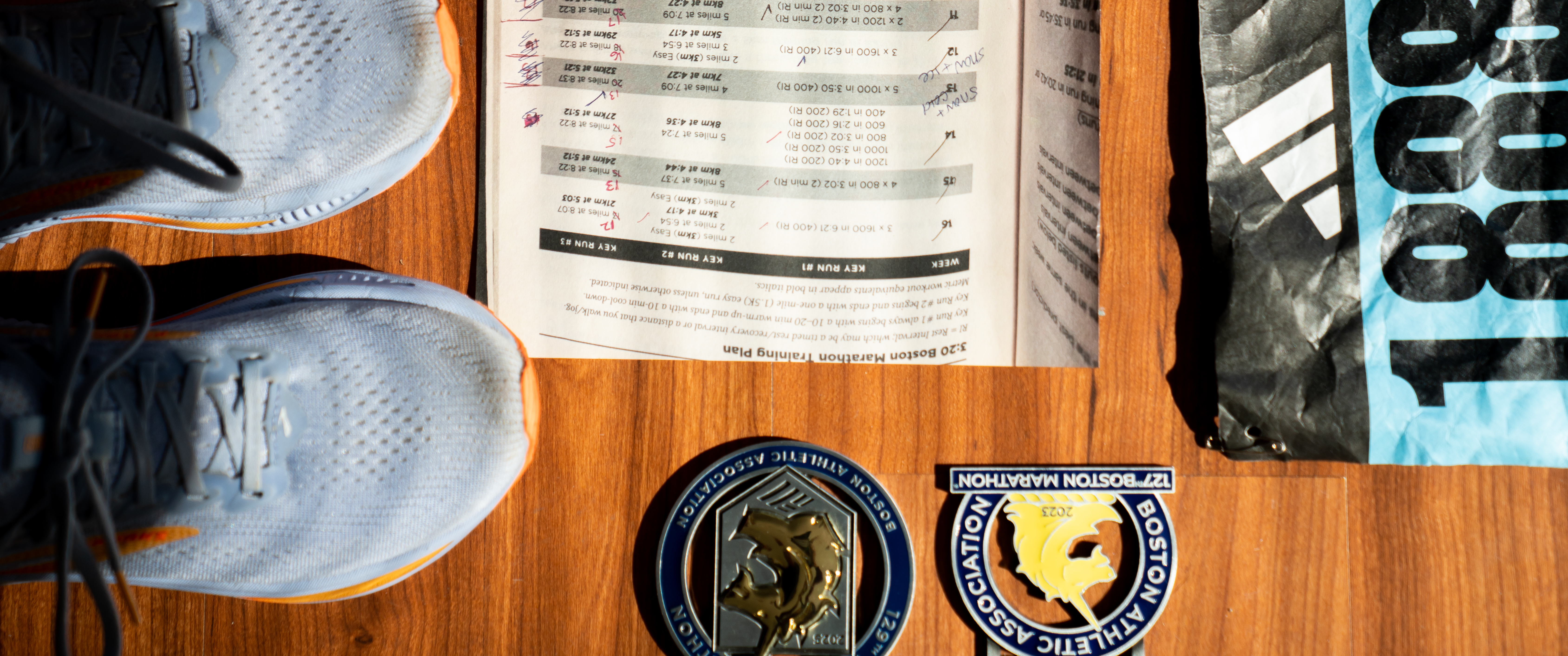 A Boston Marathon training book, sneakers, a marathon bib, and medals from the 127th and 129th Boston Marathons on a wooden surface