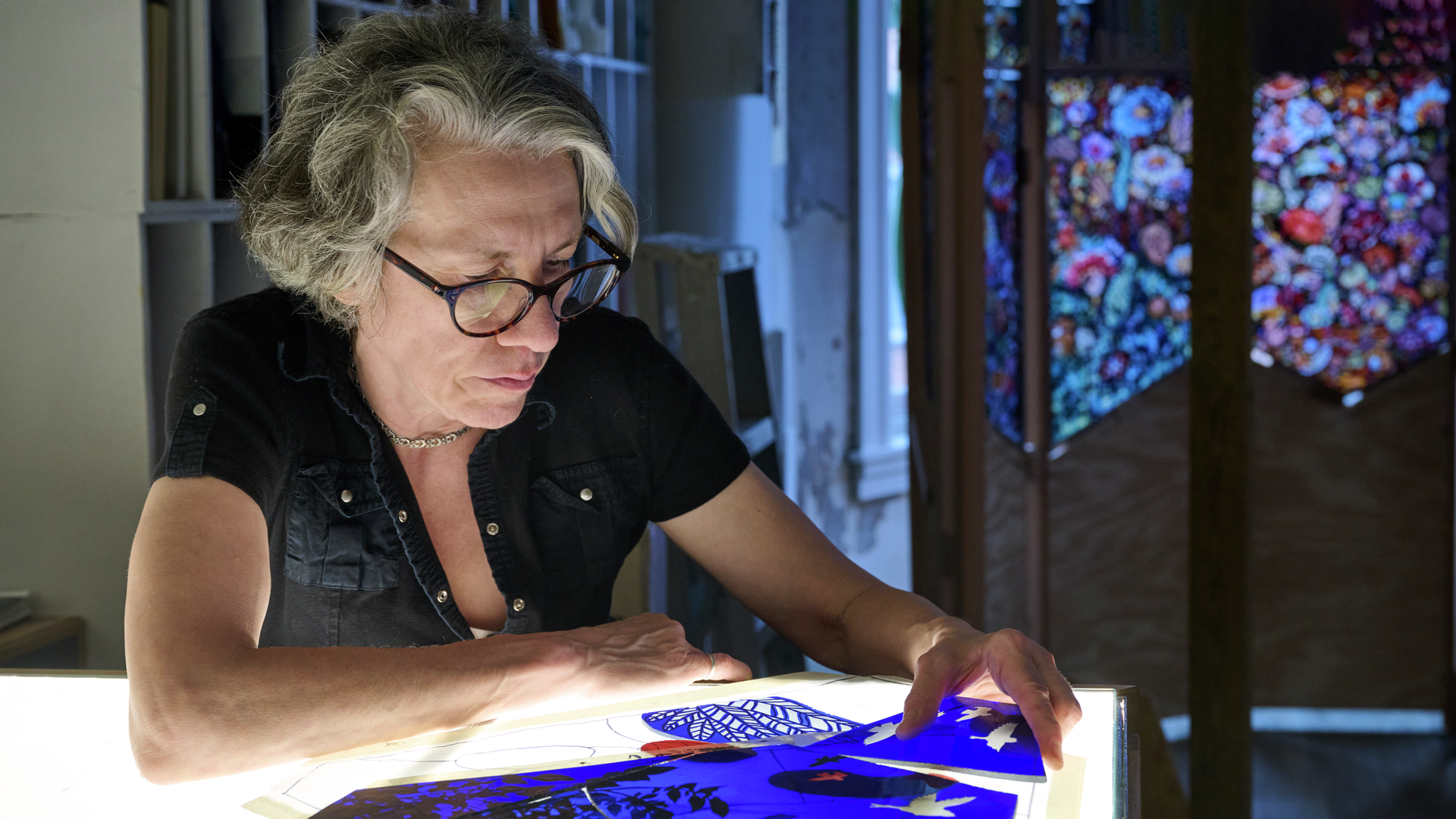 Artist Judith Schaechter sits at a table piecing together sections of glass