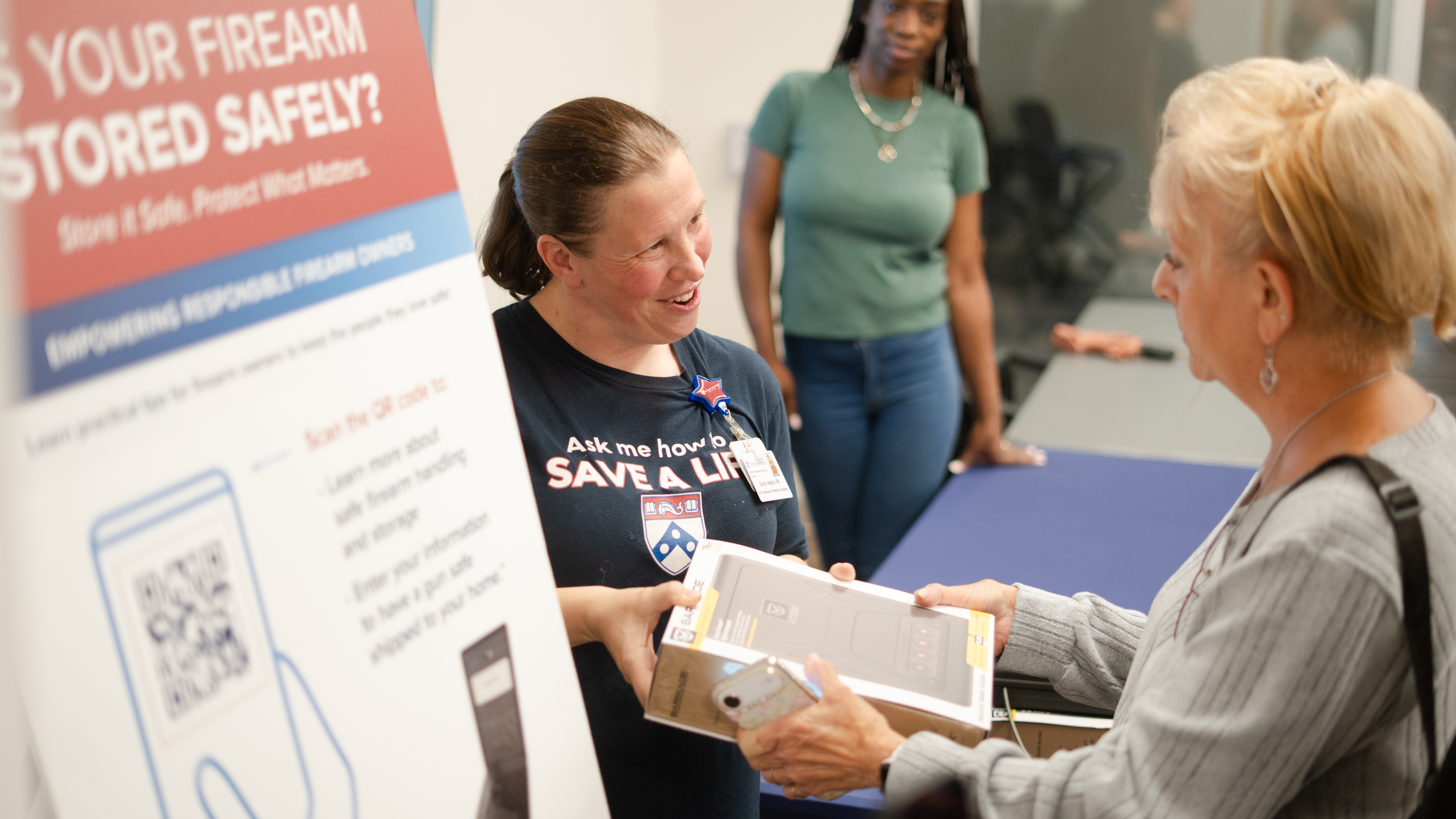 Sunny Vespico hands a woman a box containing a gun safe at a handout event in Delaware County. In the foreground is a poster displaying more information about the program.