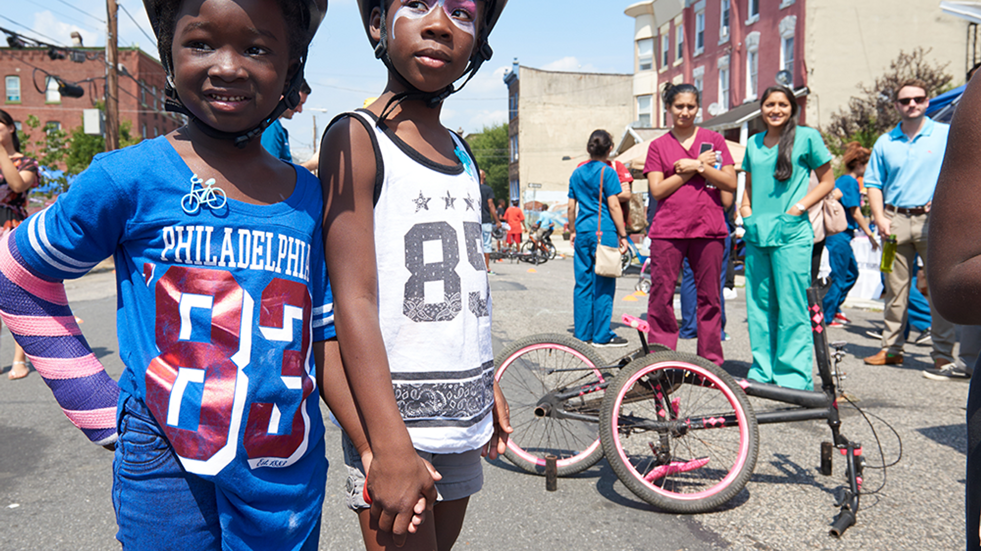 Two young boys in bike helmets hold hands at a Philadelphia street fair while health professionals in scrubs look on