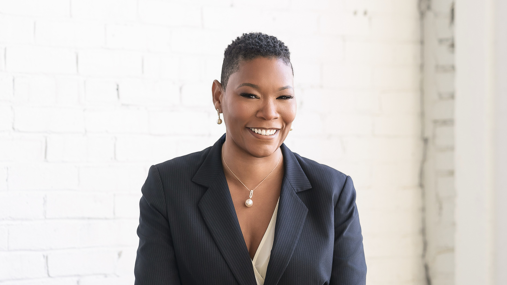 Kenya Pitt, standing in a room with a couch, smiling, wearing a pantsuit