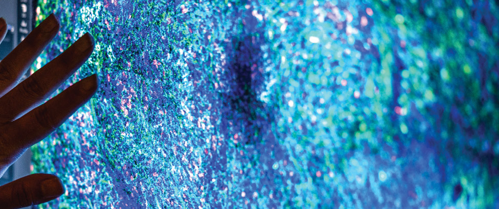 A woman’s hand is silhouetted in front of a screen showing a microscopic image of brain cells stained blue-green, with spots of pink