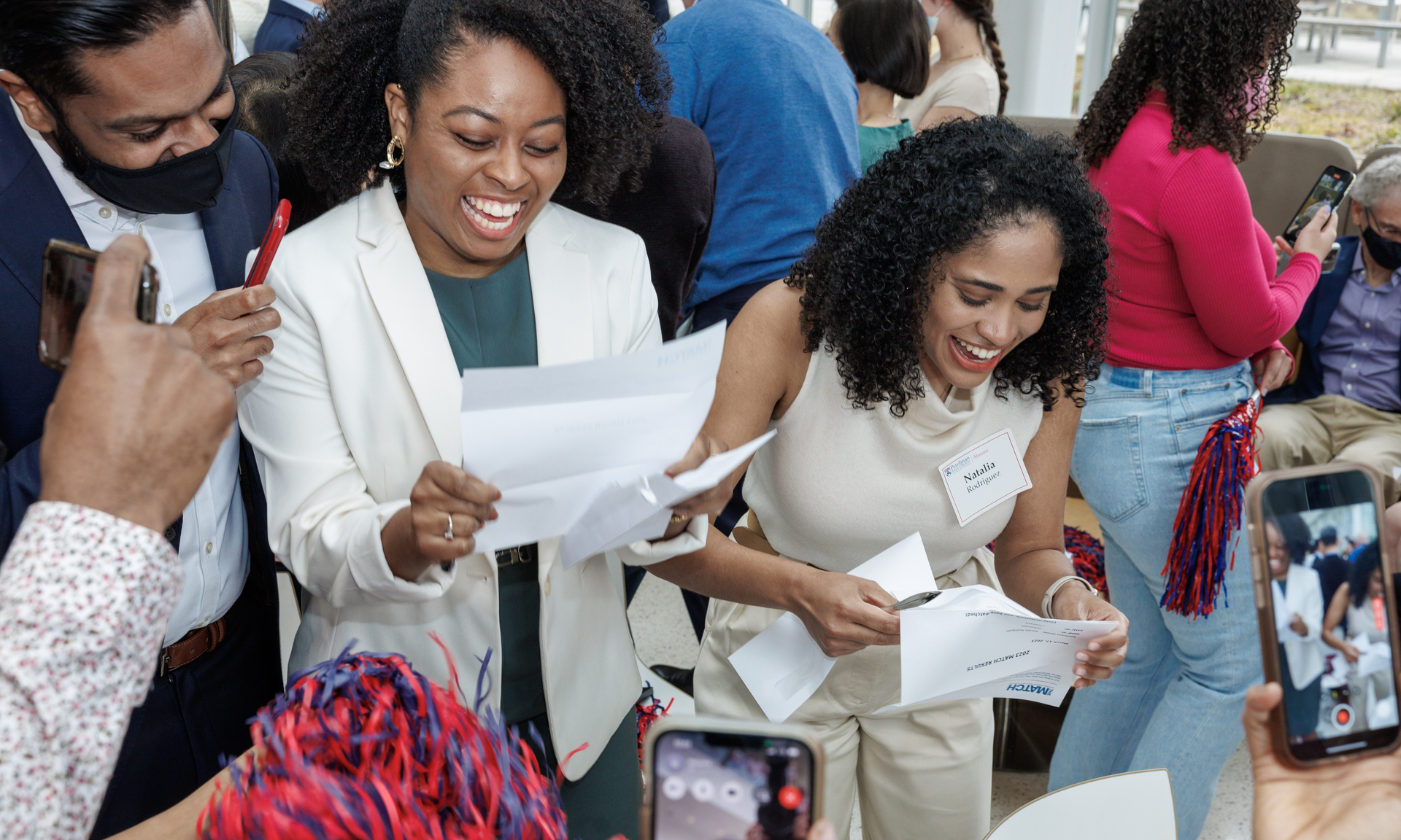 Canada Montgomery and Natalia Rodriguez stand side by side, looking at individual papers from their envelopes, with huge smiles reacting