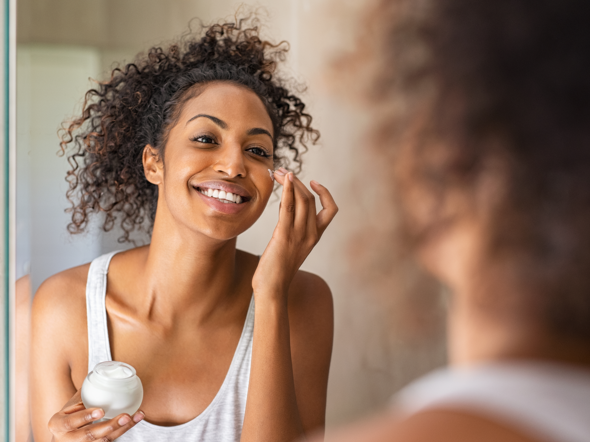Young african woman applying moisturizer on her face while standing in front of the mirror
