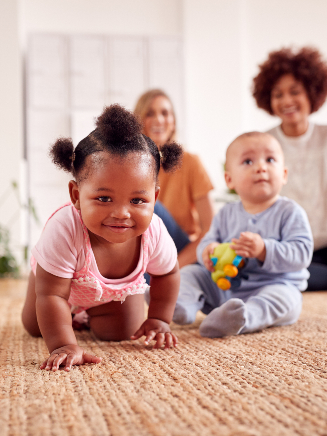 An infant crawling toward the camera on a woven rug in a bright living room, with a second infant sitting and holding a toy nearby and two adults seated in the background.