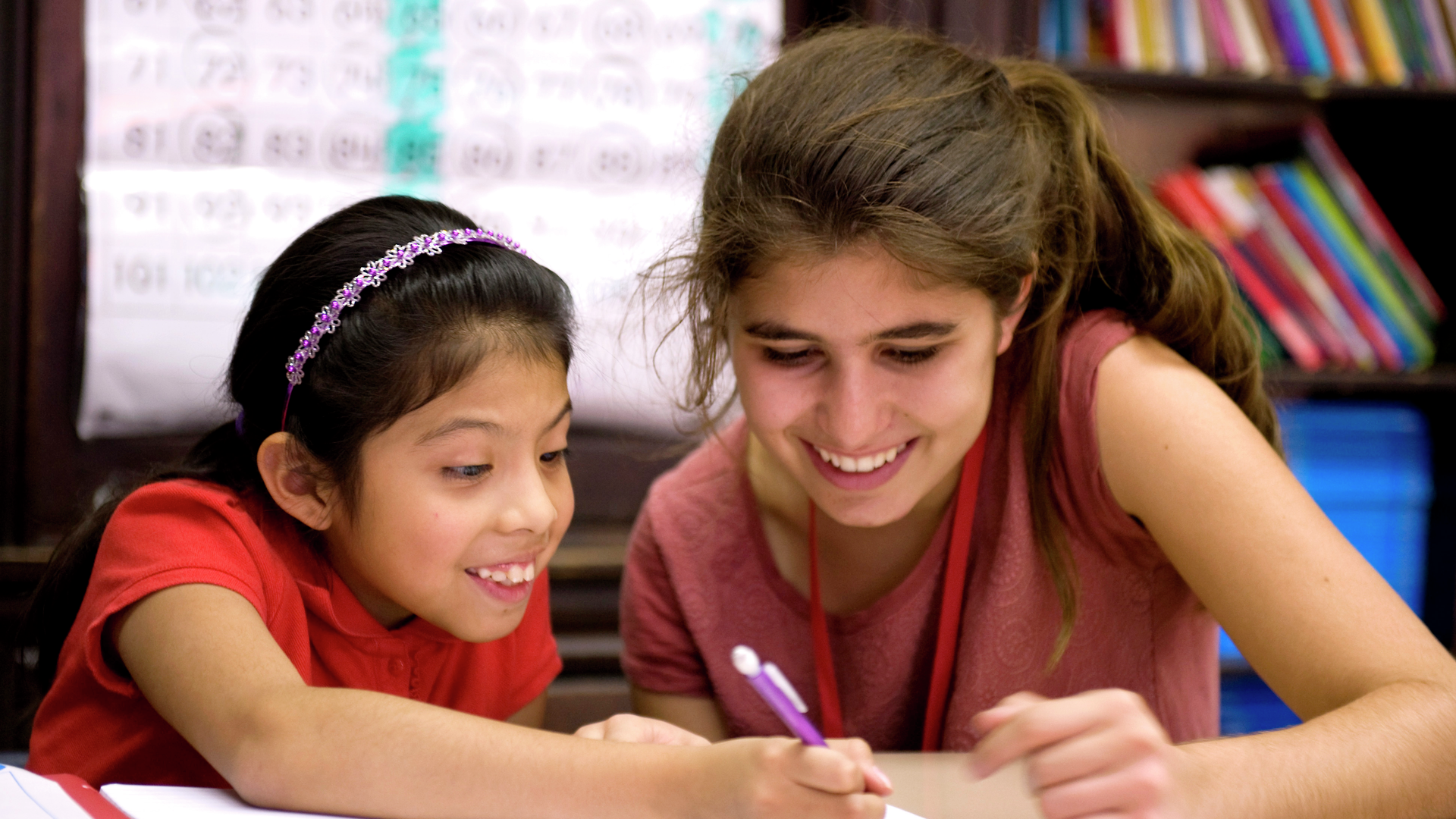 Two school-age girls smile while looking over something one of them is writing on a paper