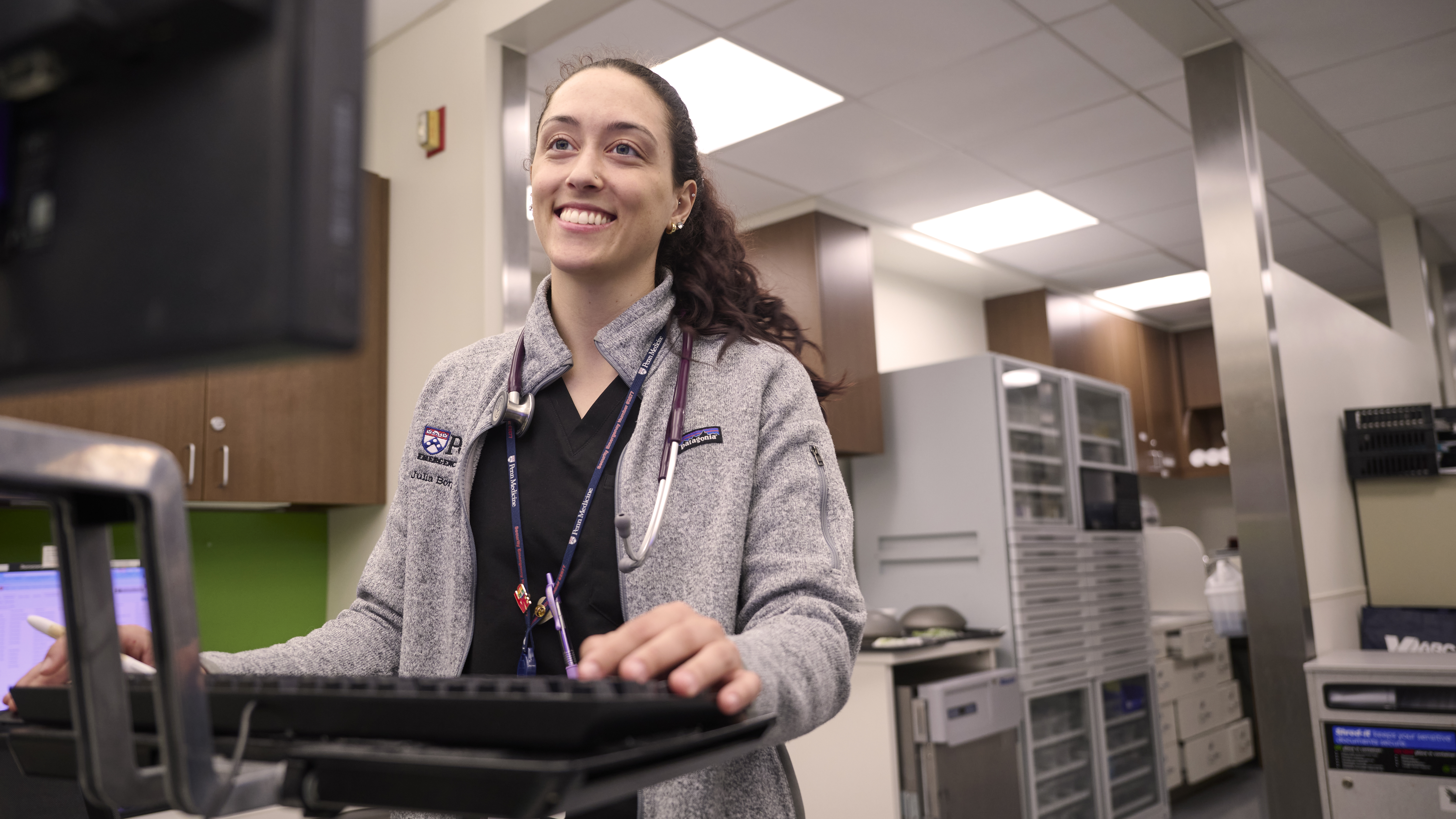 Julia Borgesi smiles while dressed in scrubs looking at a computer monitor