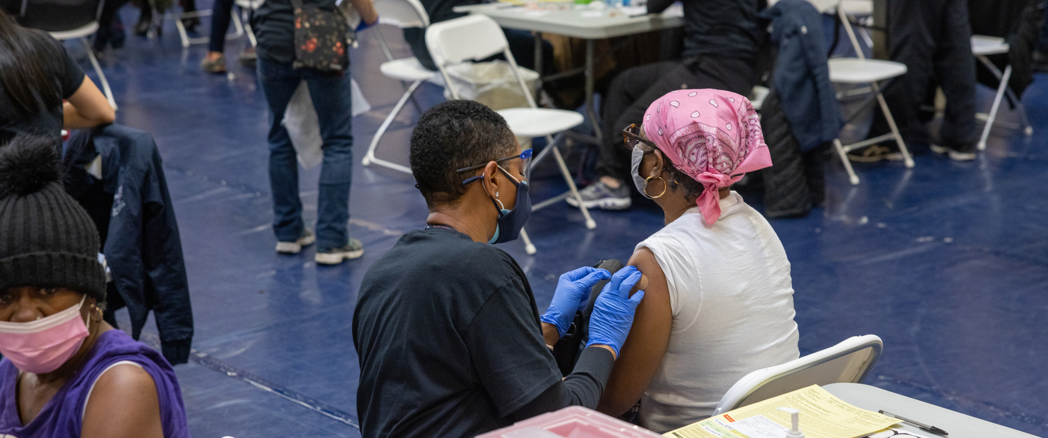 A Black female clinician administers a vaccine to a Black female patient in a school gym