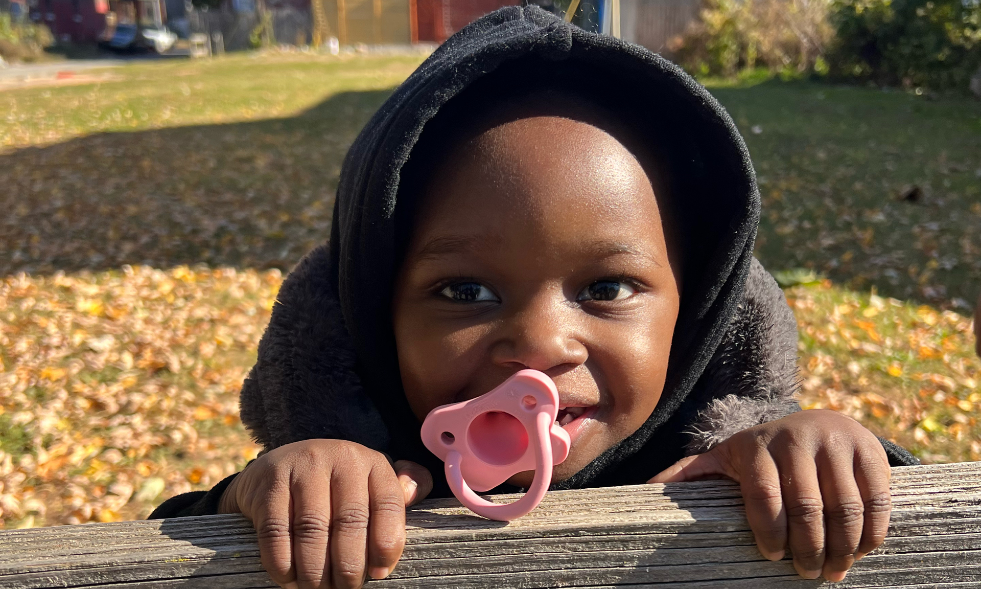 Zahiri, age 2, peeks over the edge of a fence smiling, with a pink pacifier slipping from her mouth