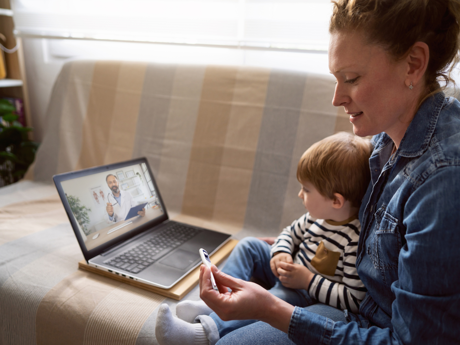 Mother with her son, having a video call with a doctor from their home while holding thermometer.