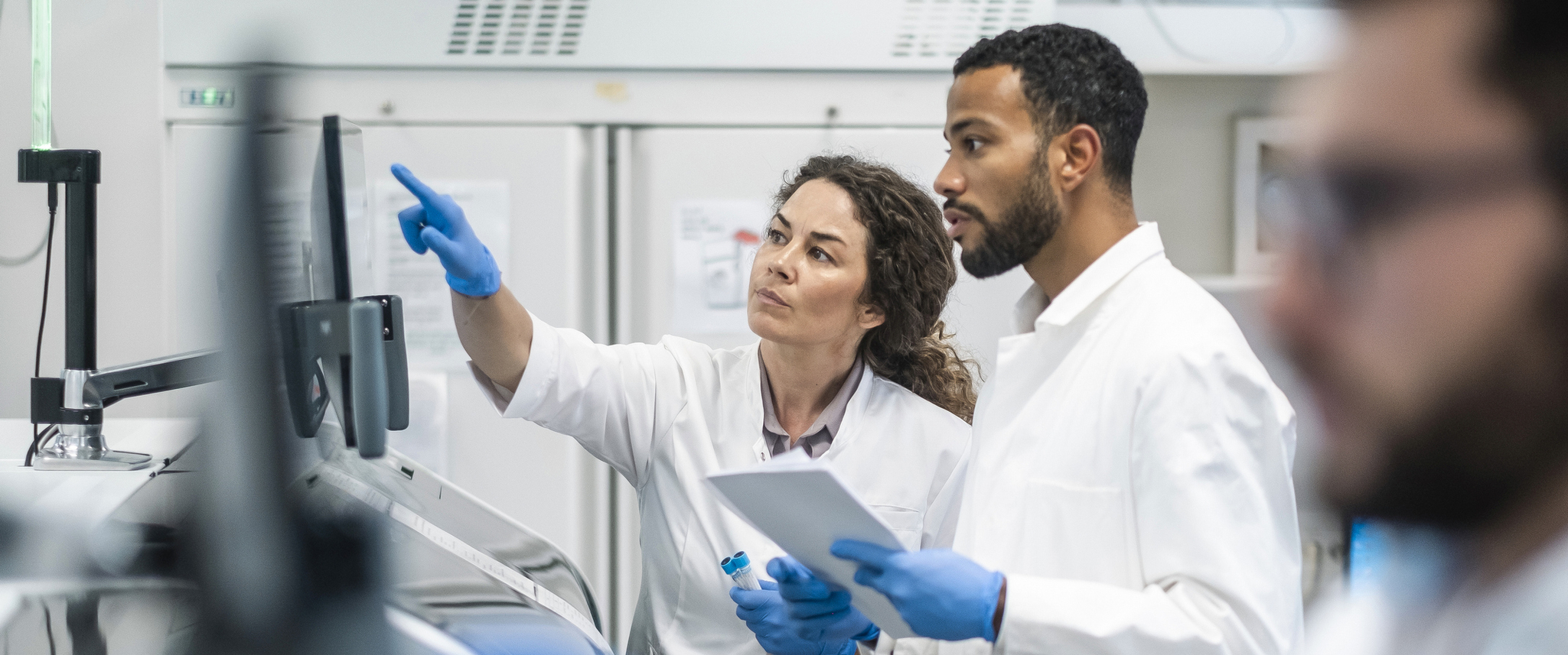 A Caucasian woman and a  man, both in lab coats, engage over research data in a bright laboratory setting.