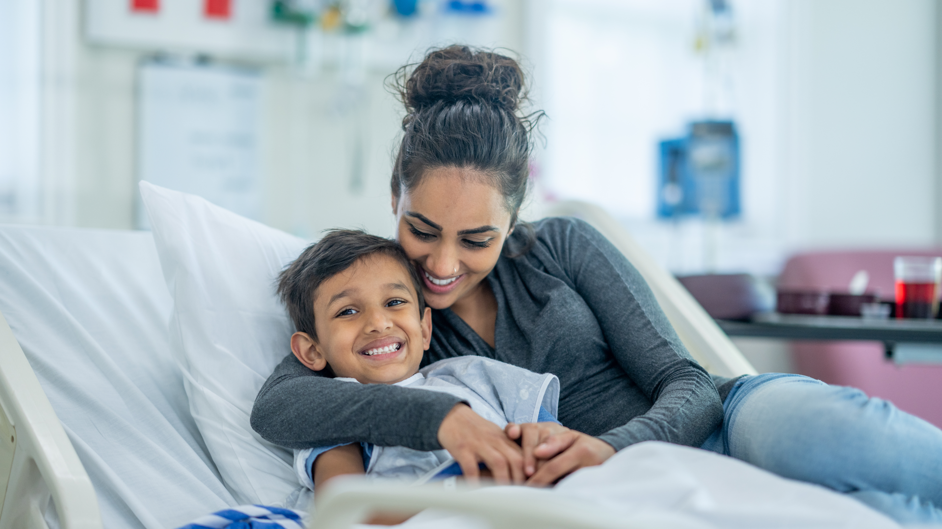 A mother sits in the hospital bed with her son as she comforts and reassures him before surgery. She is dressed casually and is puling the boy in close to her.
