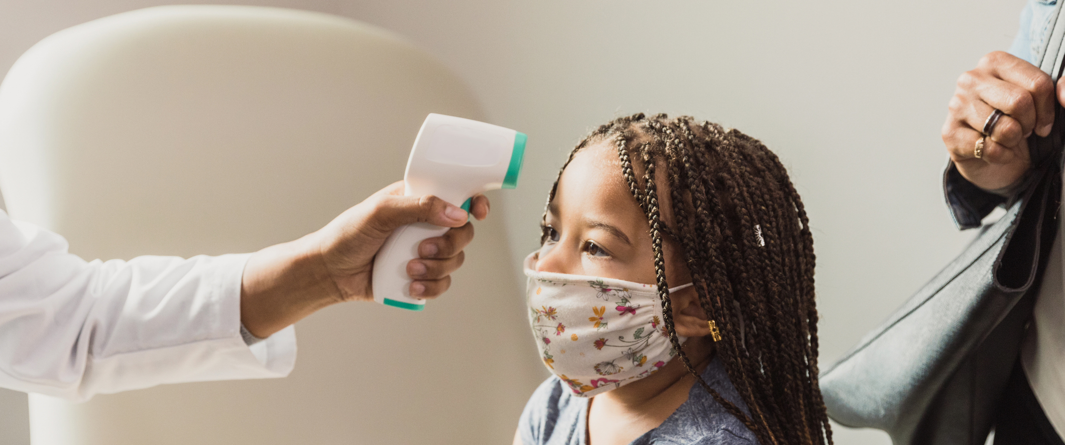A young masked girl having temperature taken by a medical professional.