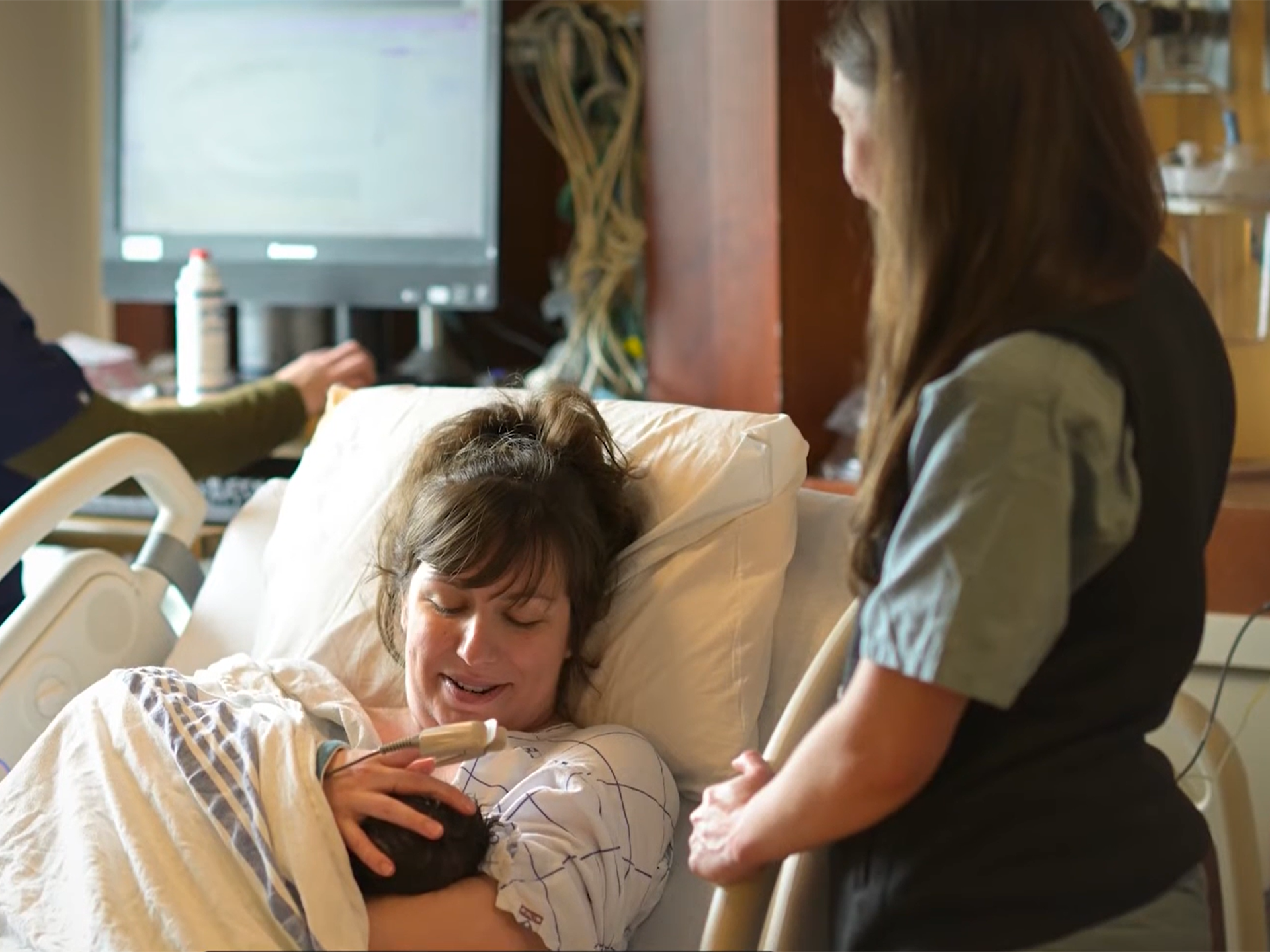 Mother and newborn in a maternity room with visitor standing by her bed