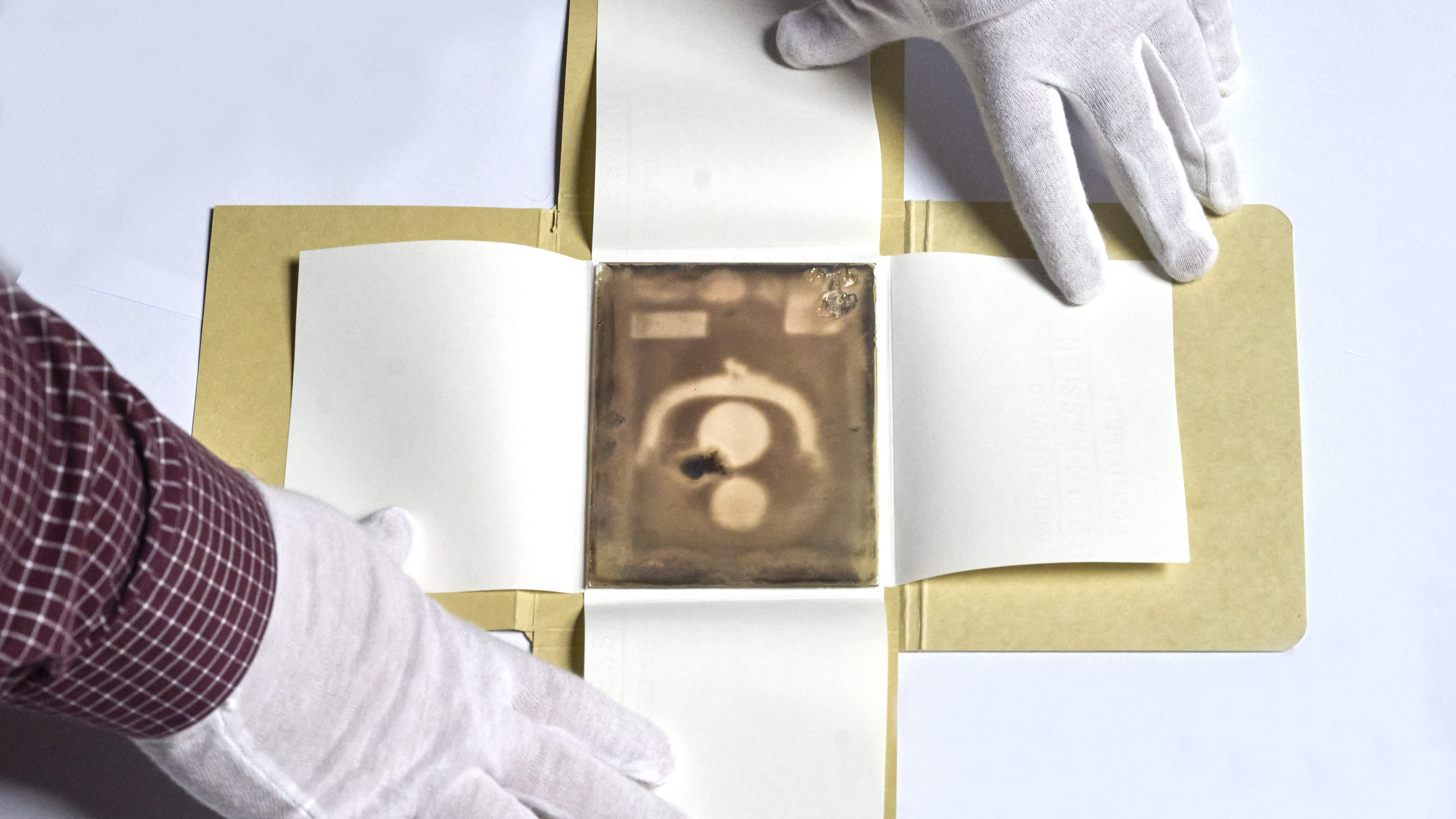 A view of a photographic glass plate showing a coin purse with two coins inside. Two gloved hands hold open the cardboard housing the glass.
