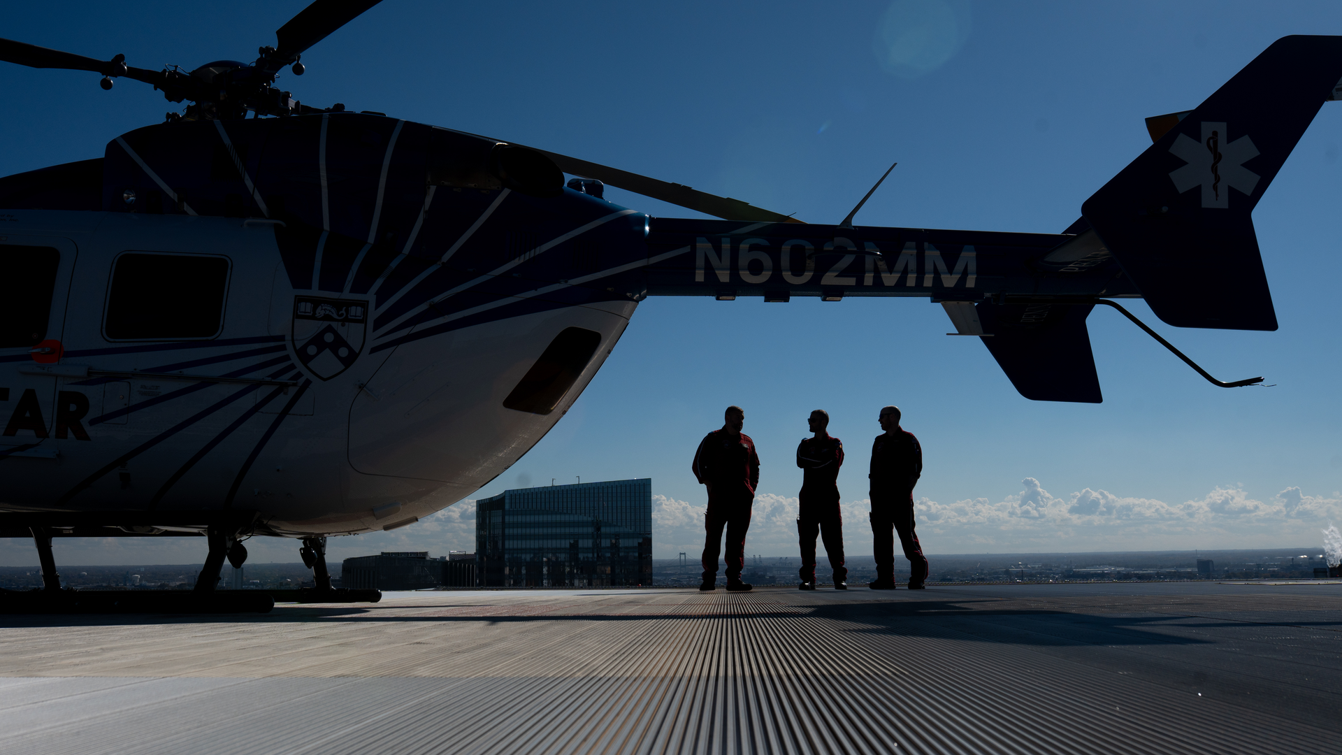 Three PennSTAR crew members stand underneath the tail of a helicopter on the rooftop of the Clifton Center silhouetted against the skyline of Philadelphia in the background