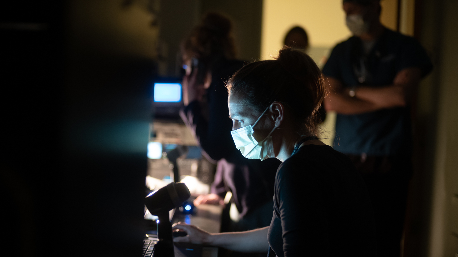 In a dimly lit production studio, a woman wearing a mask sits in front of a computer keyboard and microphone