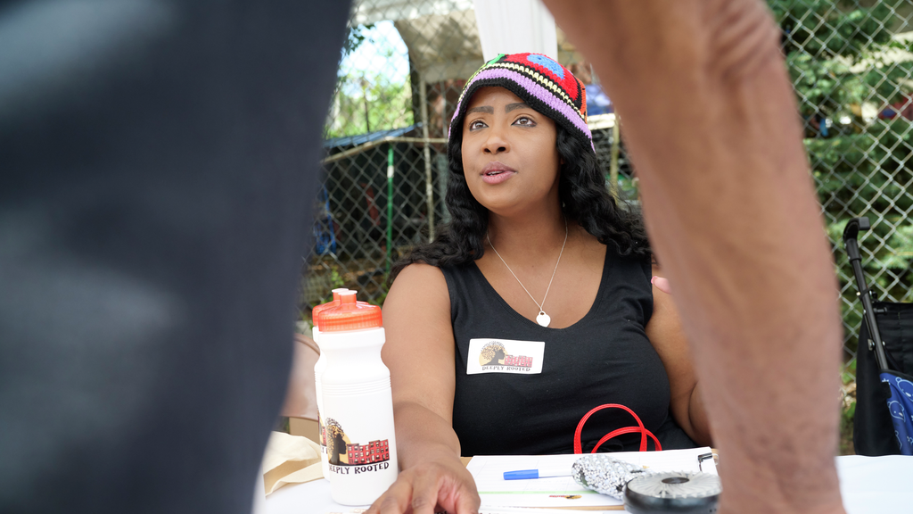 Janaiya Reason, wearing a Deeply Rooted name badge, talks with a person at a table at an outdoor community event