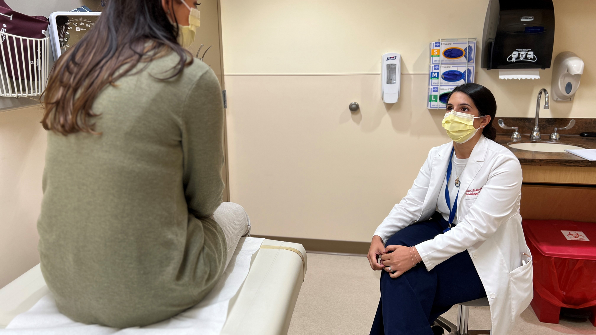 Hematologist Shivani Bakhshi, DO, at right, speaks with a woman seated on an exam table