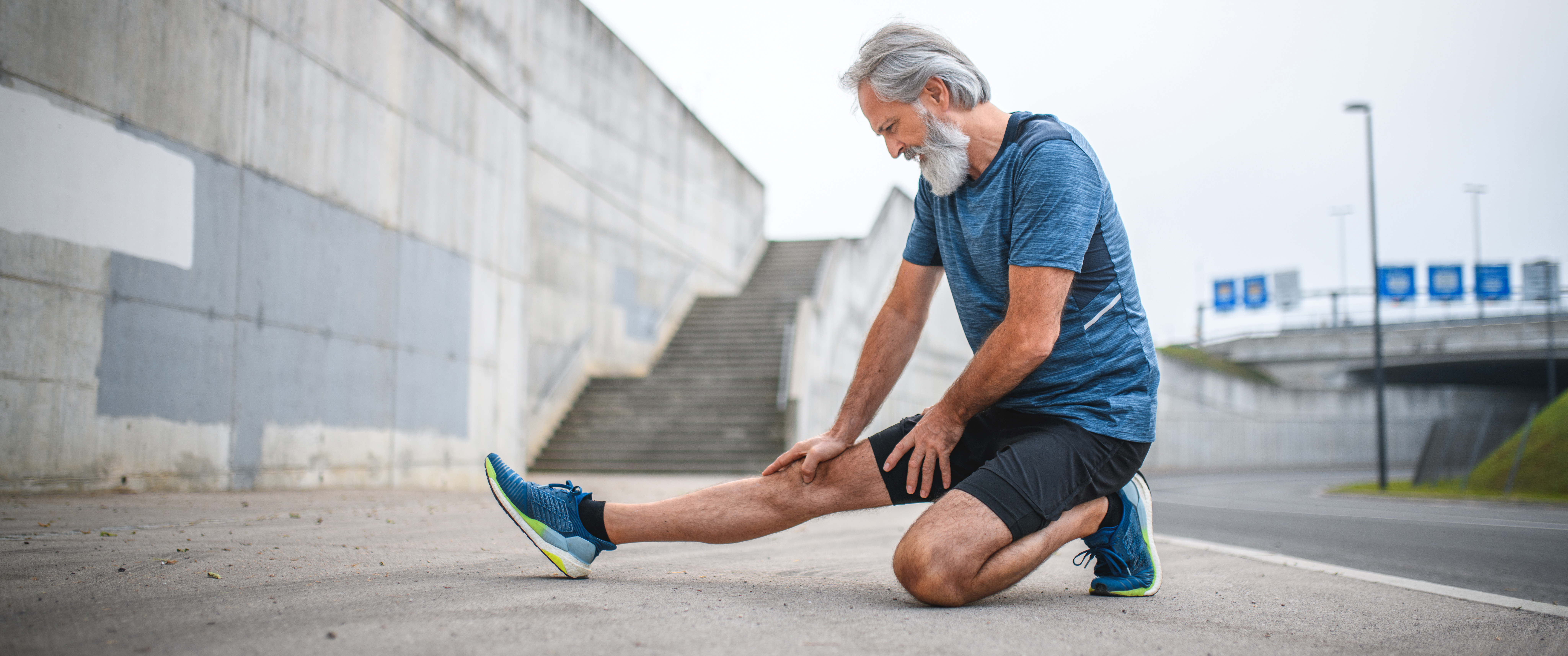 An older man kneeling outside with one leg extended out in front of him, stretching.