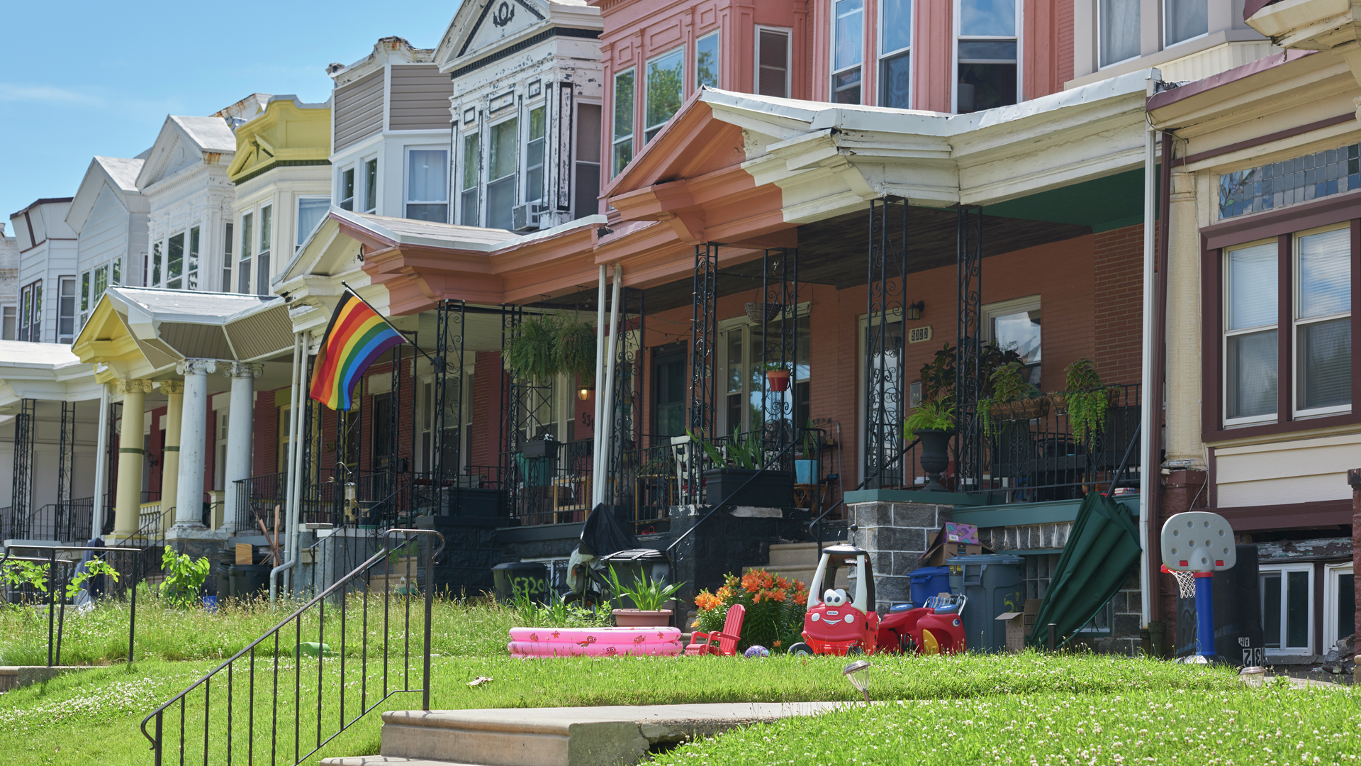 Row houses in Philadelphia of various colors, one with a Pride flag and one with children's toys on the front porch and lawn