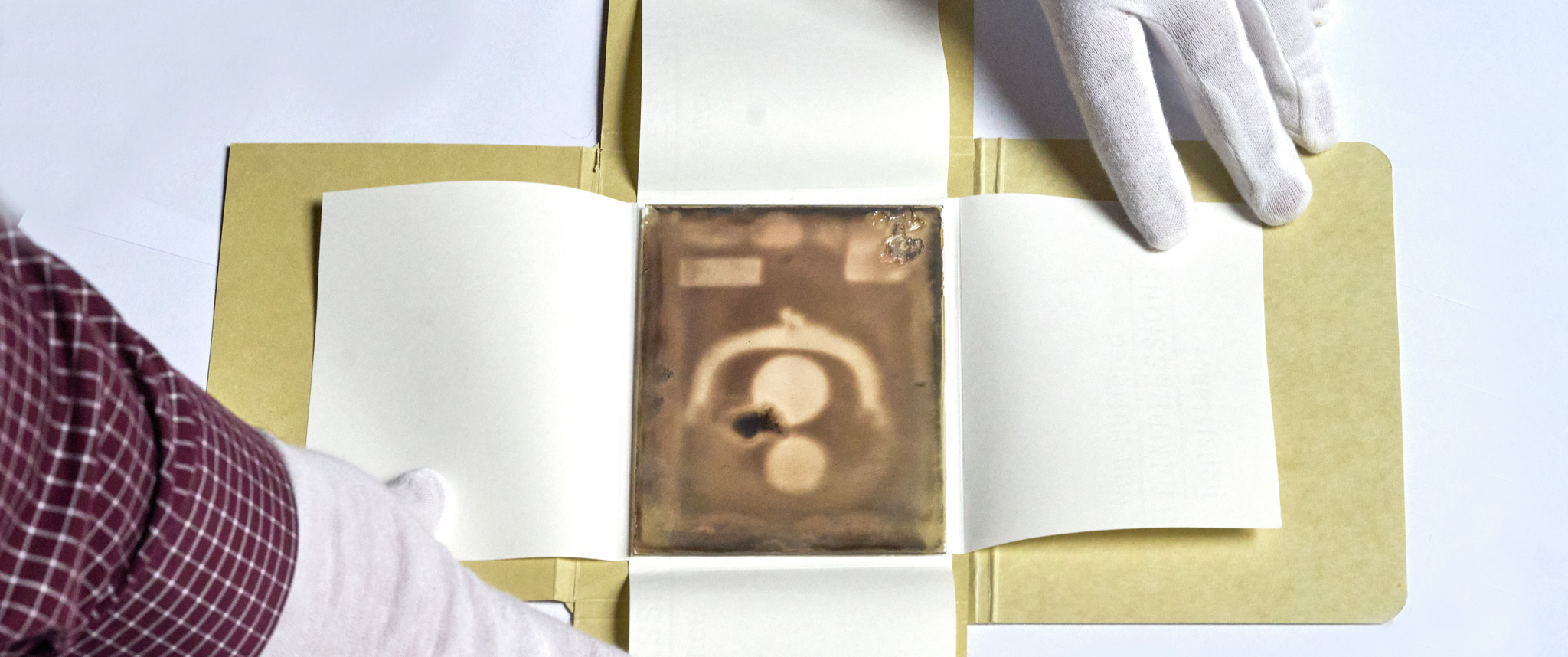 A view of a photographic glass plate showing a coin purse with two coins inside. Two gloved hands hold open the cardboard housing the glass.