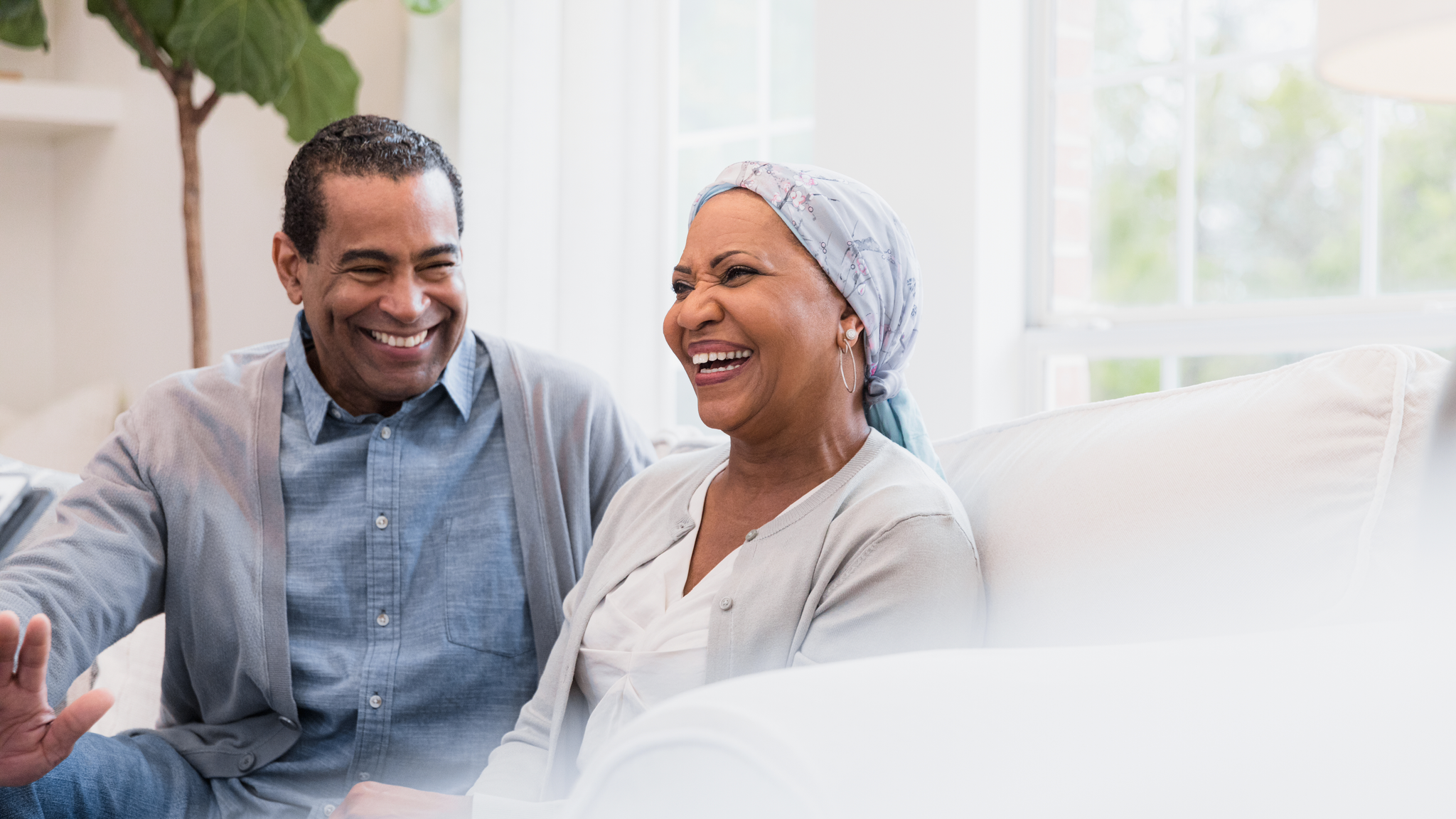 Sitting on their sofa at home, the senior adult couple smiles and laughs at a joke
