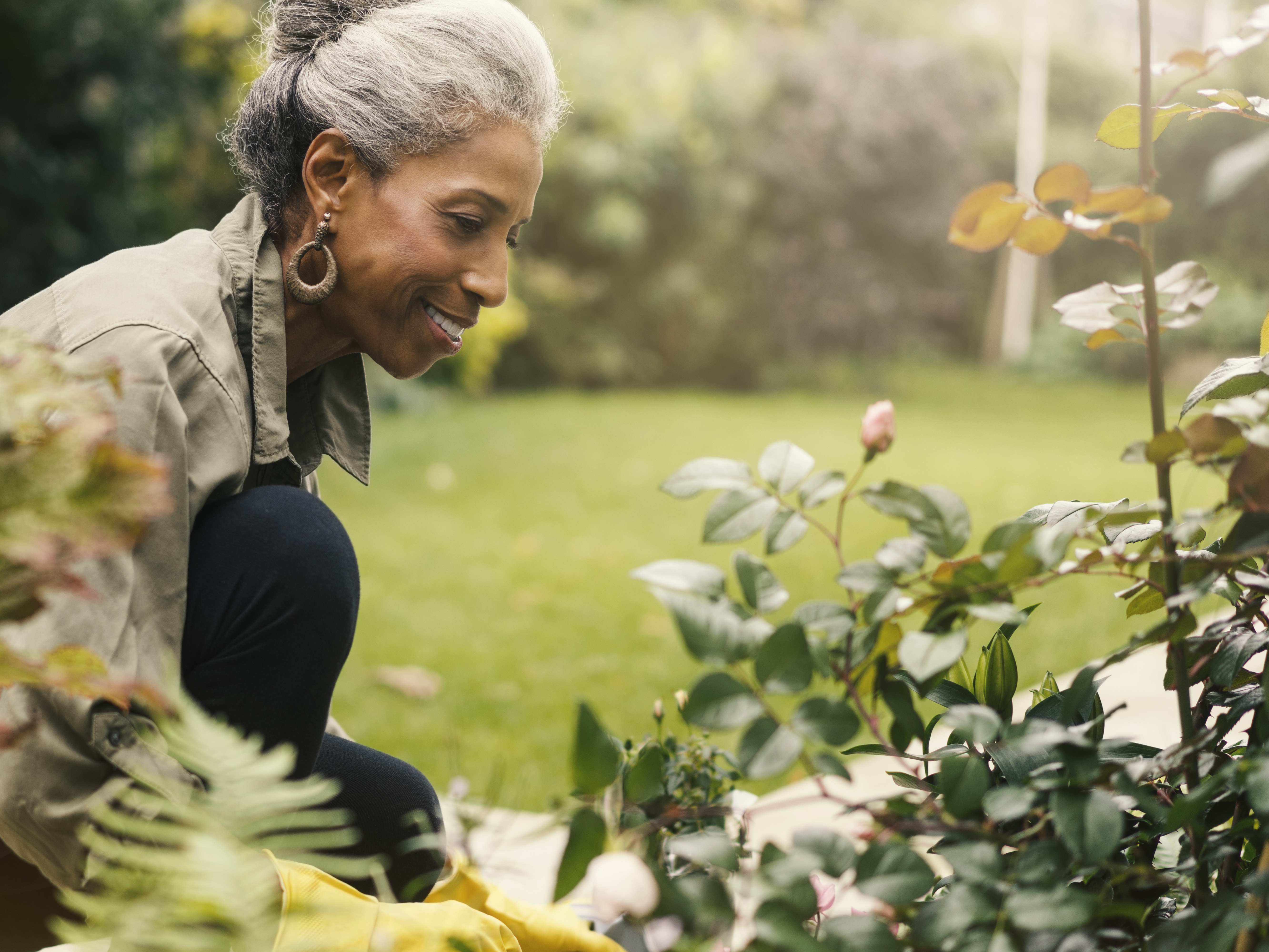 An older woman smiling while kneeling to look at a plant in her garden.