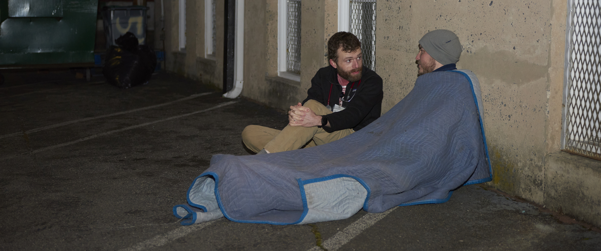 In a parking lot, a doctor sits on the ground speaking with a man lying under a blanket