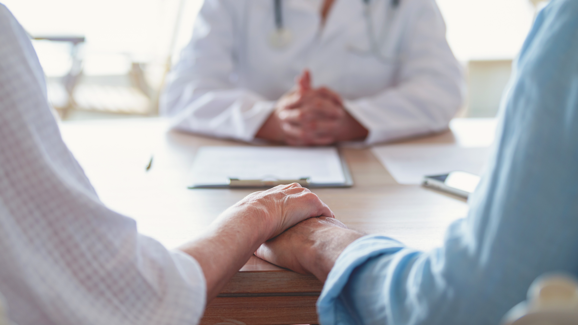 A couple is holding hands and face a doctor behind a desk with clasped hands.