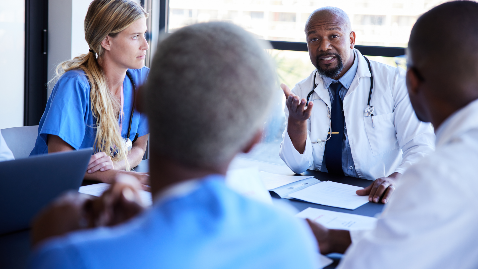 Physician talking with his diverse team of medical colleagues during a meeting around a conference table in a hospital