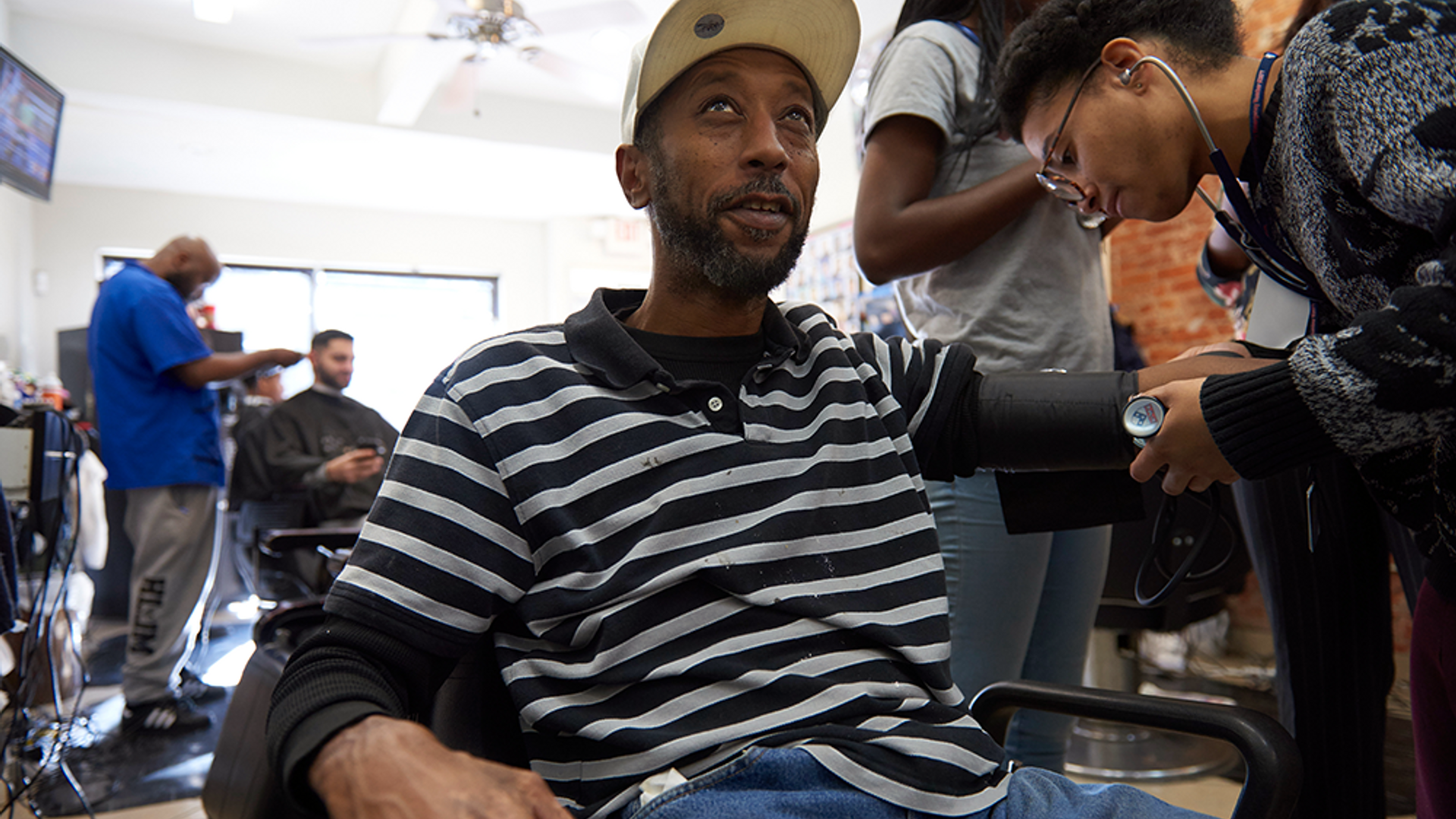 A medical student measures a Black man's blood pressure in a barbershop chair